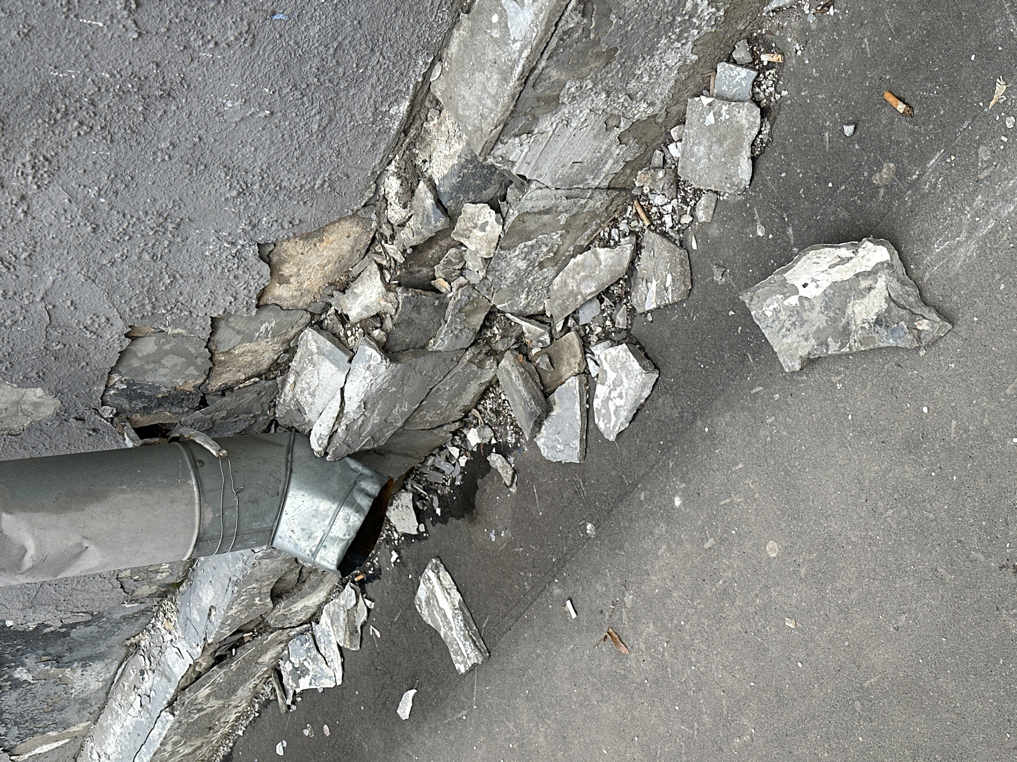 The image shows a damaged sidewalk, with a metal pipe running along the edge. The sidewalk is broken and cracked, with pieces of concrete scattered around. There is a cigarette butt on the sidewalk. It is unknown if this is in Moscow. There are no people present.