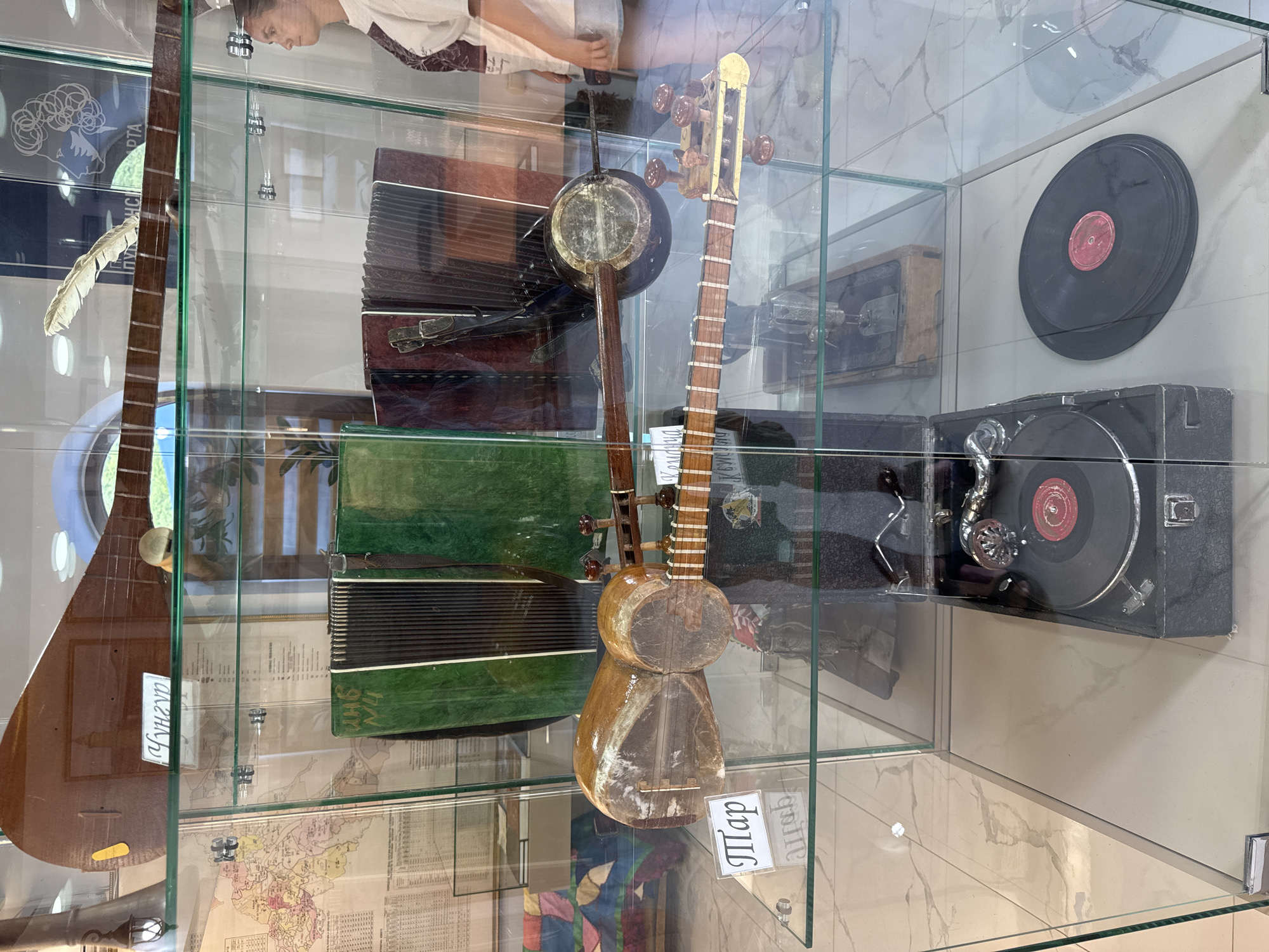 The image shows a museum display case filled with musical instruments and related items. In the top shelf, a long-necked stringed instrument is visible, along with a label. The second shelf holds two accordions, one green and one dark red, and another stringed instrument with a round body. A label is attached to this instrument. The bottom shelf features an old-fashioned record player with two vinyl records. The museum appears to be located in Khuchni, Russia, as suggested by a map visible in the background of the second shelf, which is labeled with the city name. A person is partially visible on the right side of the image, looking into the display case. The environment is indoors, with the museum exhibits presented behind glass.