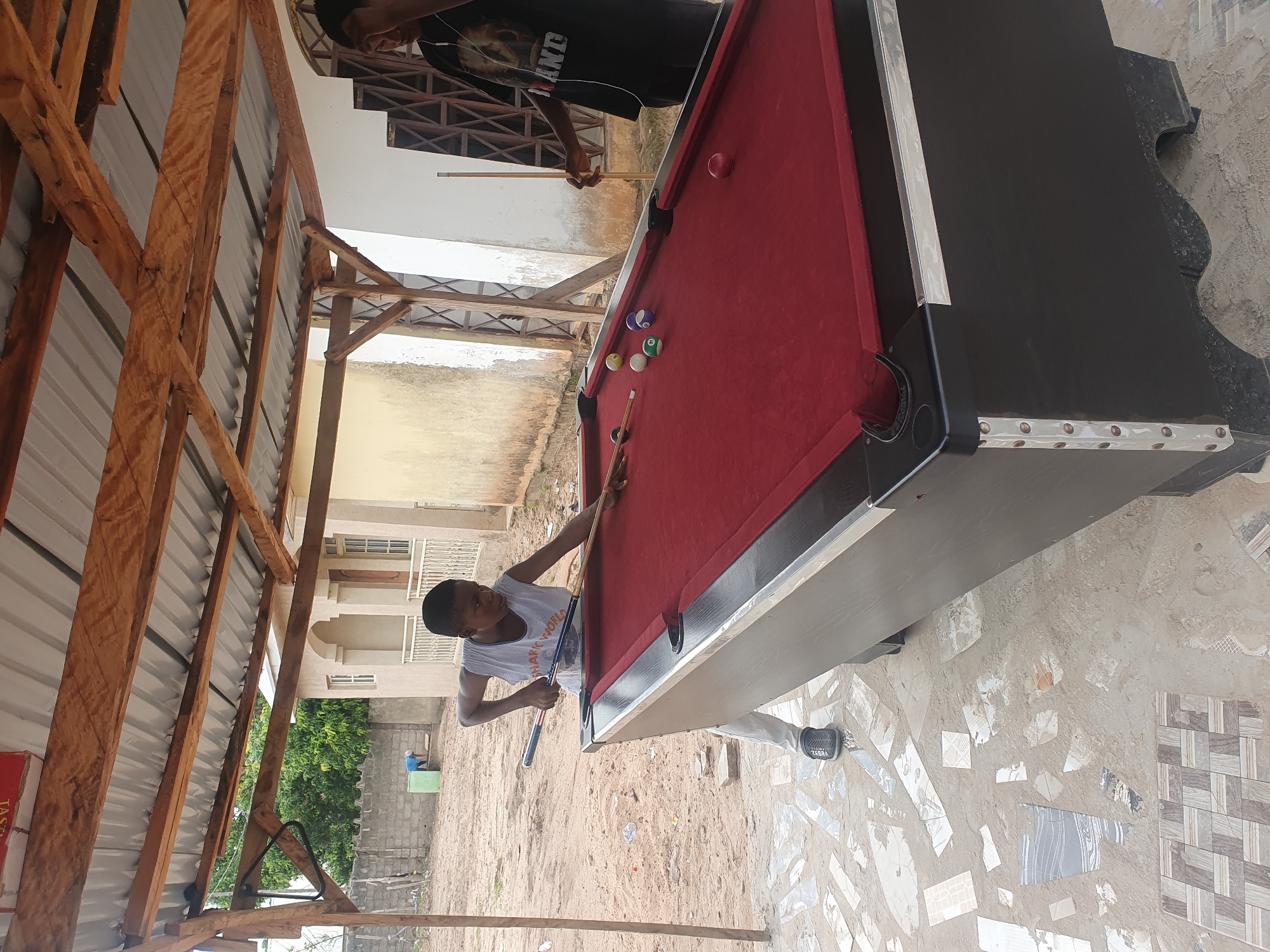 In Lokoja, Nigeria, under a simple, corrugated metal roof supported by unfinished wooden beams, a young Black boy is playing pool. He is dressed in a white tank top with the words "SHARK WORKS" printed on it and light-colored pants. He holds a pool cue, poised to strike a white ball on the red felt of the pool table. Several other colored billiard balls are scattered on the table. To the right, another Black individual, wearing a black t-shirt and headphones, stands near the table, observing. The environment appears to be outdoors, with sandy ground and scattered debris, including pieces of broken tiles on the pavement around the pool table. In the background, a light-colored building with arched windows and a white railing is visible, along with a grey cinder block wall and some trees.
