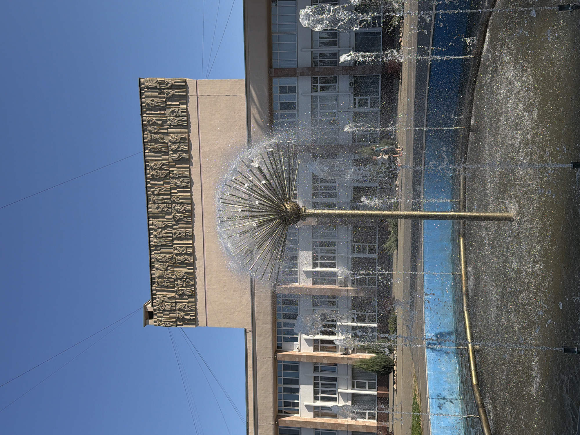 This image depicts a building in Volzhsky, Russia, adorned with a bas-relief frieze along its upper facade, a common architectural feature in Soviet-era public buildings. In the foreground, a fountain sprays water, creating a dynamic visual element that partially obscures the building's facade. The building itself is a multi-story structure with numerous windows, suggesting it might be an administrative or cultural institution. The sky is clear and blue, indicating fair weather. Two people are visible walking on a paved area in front of the building, to the left of the fountain. The overall scene suggests an urban public space with elements of both architecture and natural water features.