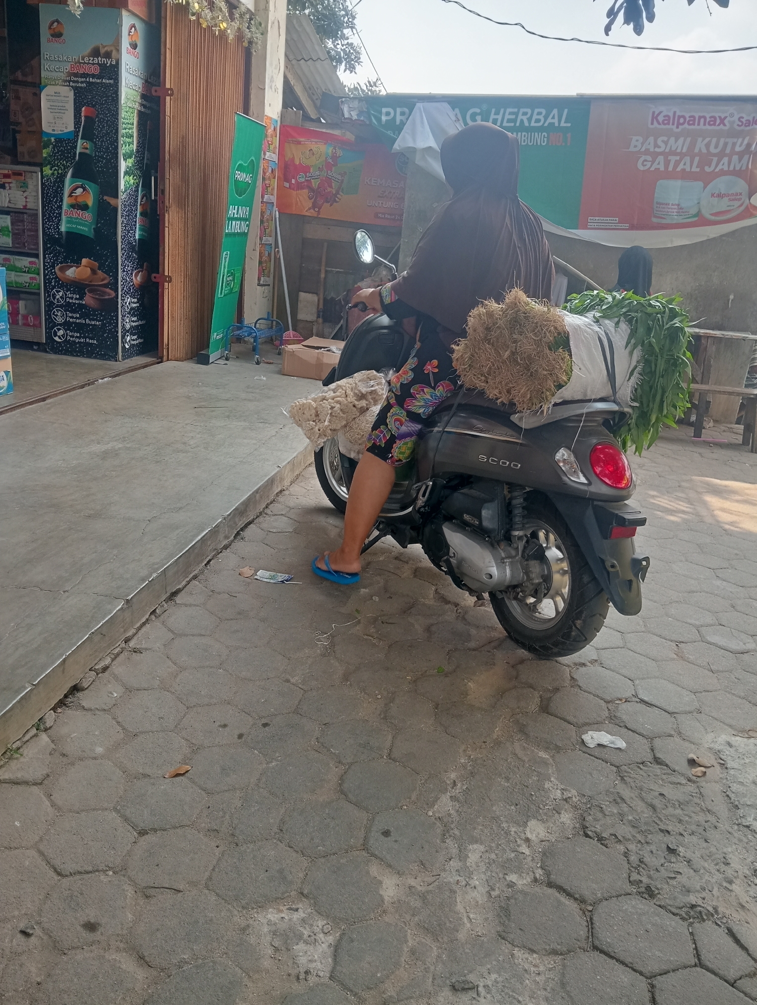 A woman wearing a brown headscarf and a floral-patterned skirt sits on a dark grey scooter. She is facing away from the camera. The scooter is parked on a textured concrete pavement, and a pile of dried grass or straw is strapped to the back of the scooter, along with a bunch of green leafy vegetables. In front of the woman, a bag of snacks, possibly crackers, is visible. The background shows a shop with an open sliding door, and various advertisements and posters are displayed. One poster on the left features a bottle of soy sauce and text in Indonesian. Another large banner in the background advertises "PRAG HERBAL" and "Kalpanax Salep Basmi Kutu Gatal Jamur." The environment appears to be an outdoor commercial area. The scene is illuminated by natural daylight, casting some shadows.
