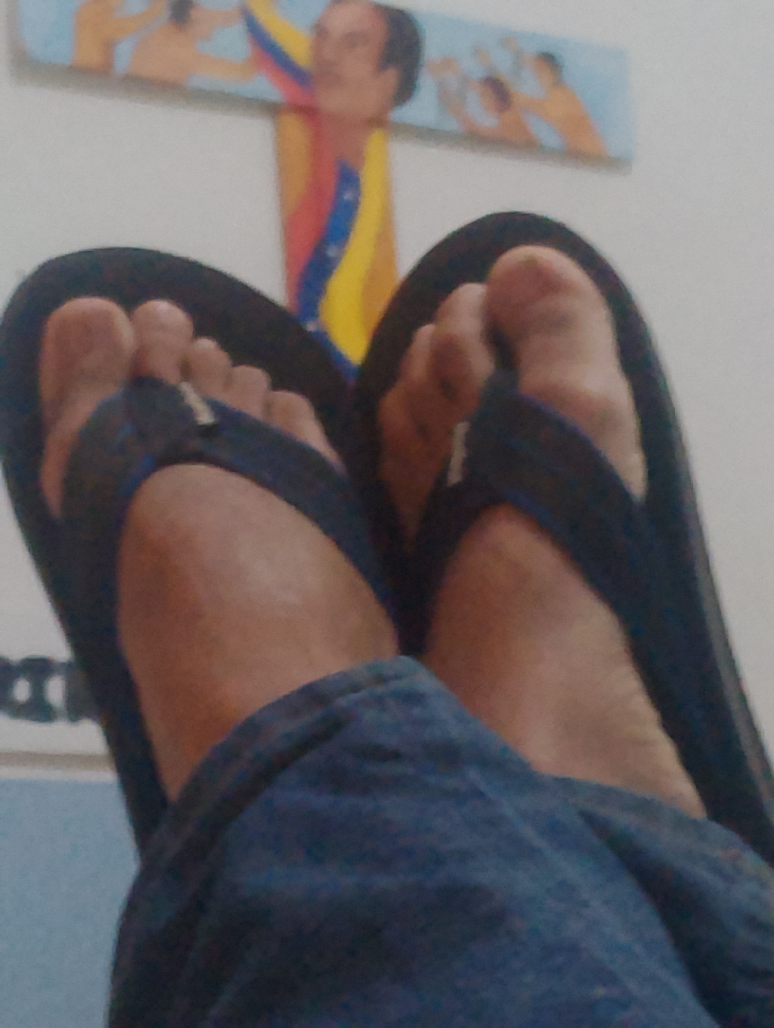A close-up shot from a first-person perspective shows a person's feet in black flip-flops with blue straps. The person is wearing blue clothing. Behind the feet, on a wall, is a piece of art featuring figures, including one of Hugo Chavez draped with the Venezuelan flag. There is no indication in the image that the location is Pampatar, Venezuela.