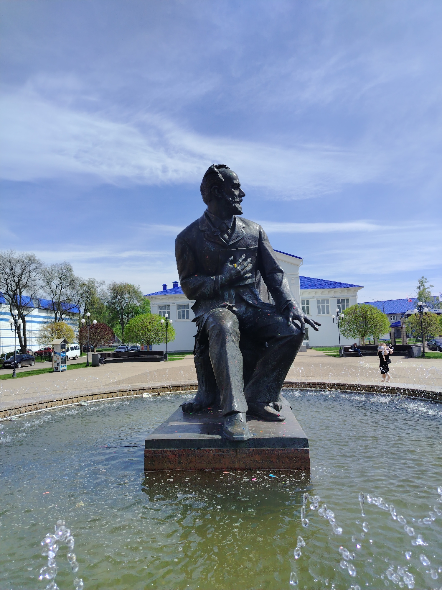 The image shows a bronze statue of a seated man in a suit, placed on a stone pedestal surrounded by a fountain in the city of Chaikovsky, Russia. The man appears to be looking to the right, with his right hand on his chest. Behind the statue is a paved public square, with benches and trees. In the background are buildings with blue roofs and white walls. There is a child standing by the fountain's edge. Cars and a van are parked on the periphery. The sky is blue with scattered clouds.