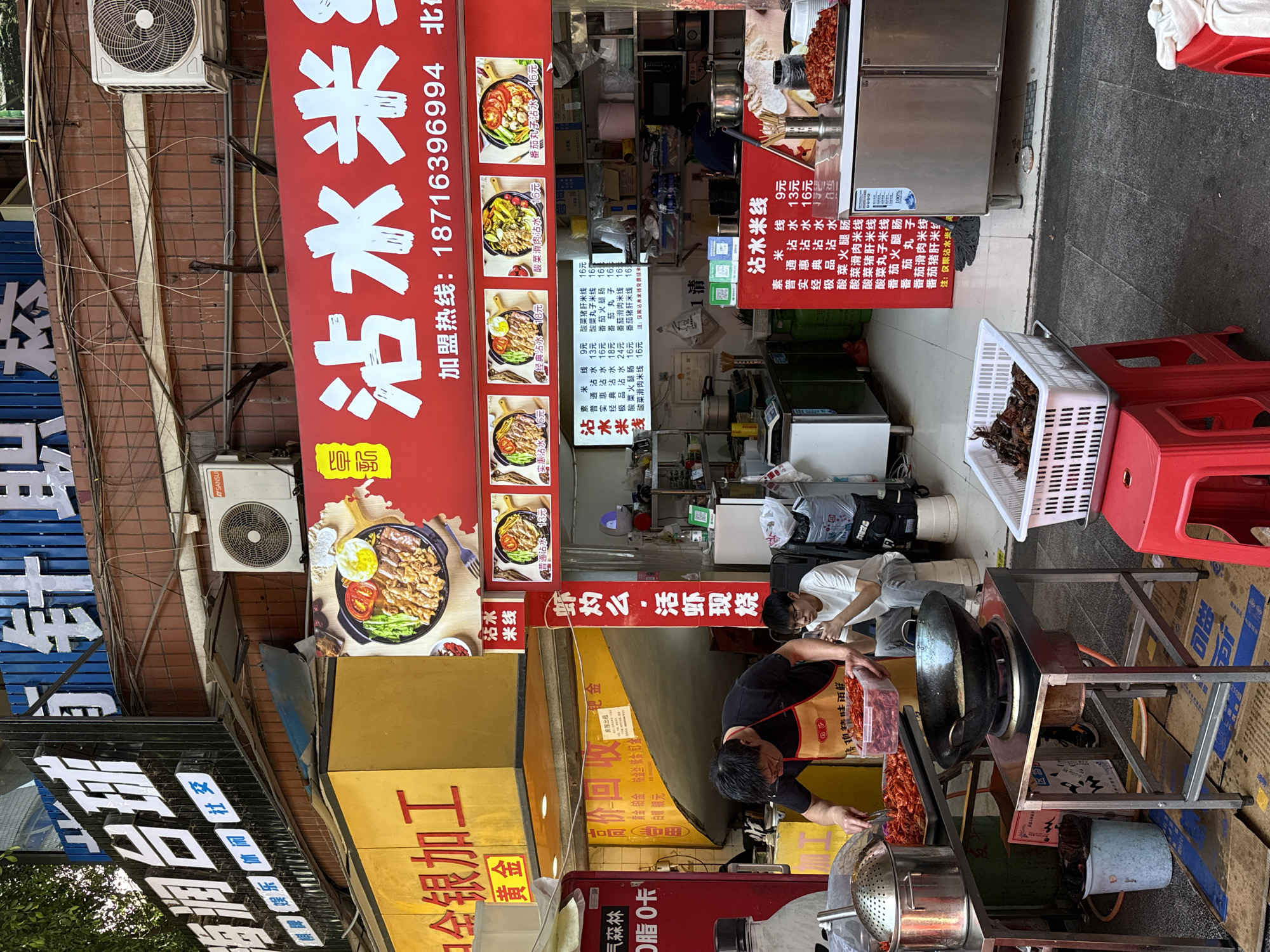 Here's a forensic description of the image:
**Location:** Beibei, China
**Scene:** A street-side food stall. The stall is serving "Zhan Shui Mi Xian" (沾水米线), which translates to "dipping rice noodles".
**People:**
* Two men are visible; one appears to be cooking with a large wok. He wears a black shirt and an apron. The other is in the background wearing a white shirt.
**Objects:**
* A large wok on a stand is visible, with flames underneath.
* A basket containing what appear to be crawfish is set next to red plastic stools.
* Several signs advertise the food being sold, with the name of the dish written in large characters. Menu items with prices are listed.
* Various kitchen utensils, including pots, and a stainless steel refrigerator are visible.
* Air conditioning units are mounted on the building behind the food stall.
**Actions:**
* The man is actively cooking using a large wok over a heat source.
**Environment:**
* The surrounding area appears to be a commercial district with various storefronts.
* The food stall is located on the sidewalk.
* The image is taken in daylight.