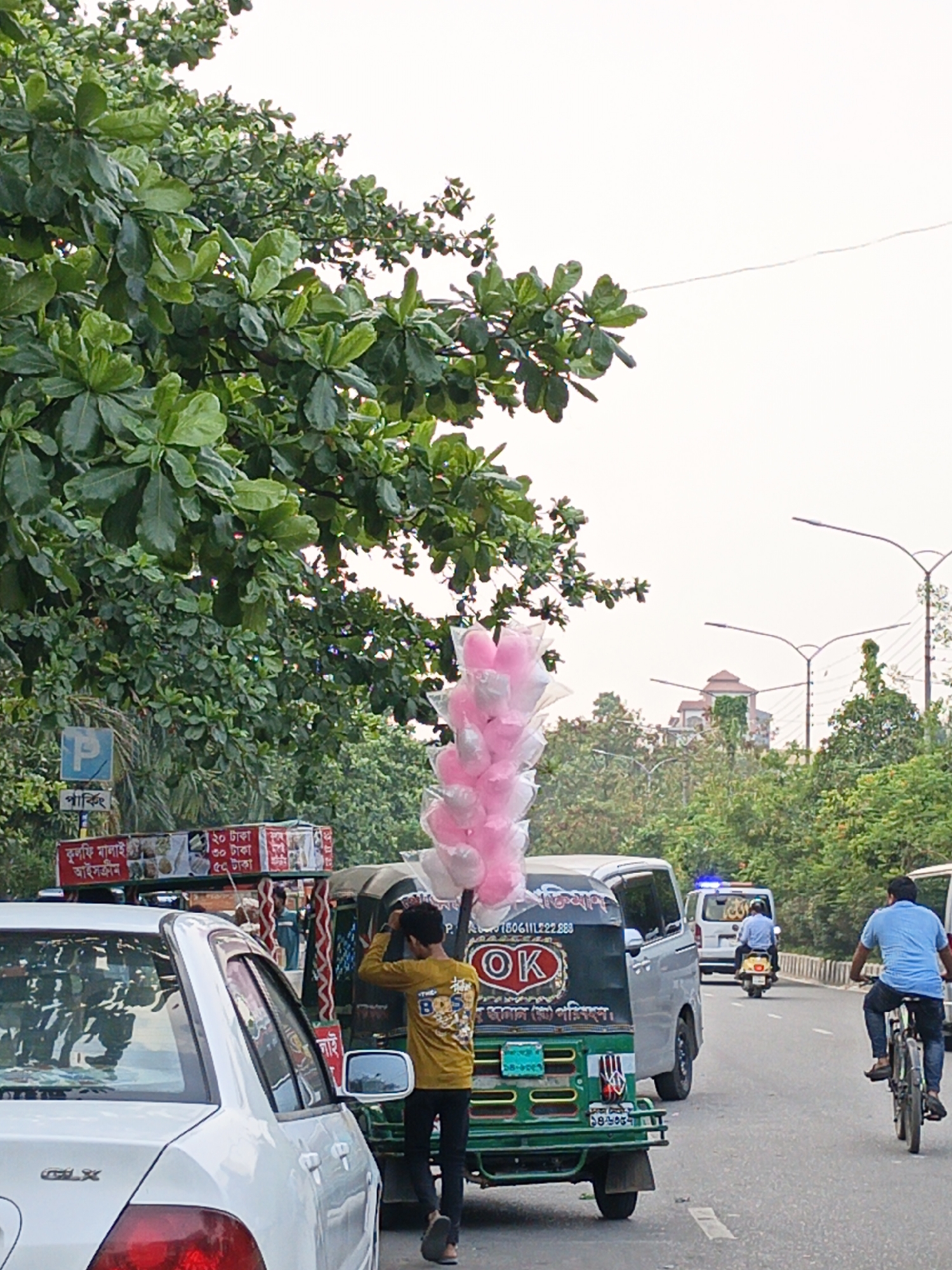 The image captures a vibrant street scene in Dhaka, Bangladesh. A roadside vendor stands near a green and black CNG auto-rickshaw, adjusting what appears to be a bundle of pink and white cotton candy or balloons atop the vehicle. To the left of the auto-rickshaw is a white sedan. Another food cart with an advertisement board for "Kulfi Malai Ice Cream" and pricing details is adjacent to a blue parking sign.
The street scene extends into the distance, revealing a gray van, a man riding a scooter with a blue siren, and a man on a bicycle. The road is lined with lush trees, providing a green backdrop. Street lights are visible overhead, and in the far distance, a building can be seen. The sky is overcast, contributing to a soft, diffused light in the scene.