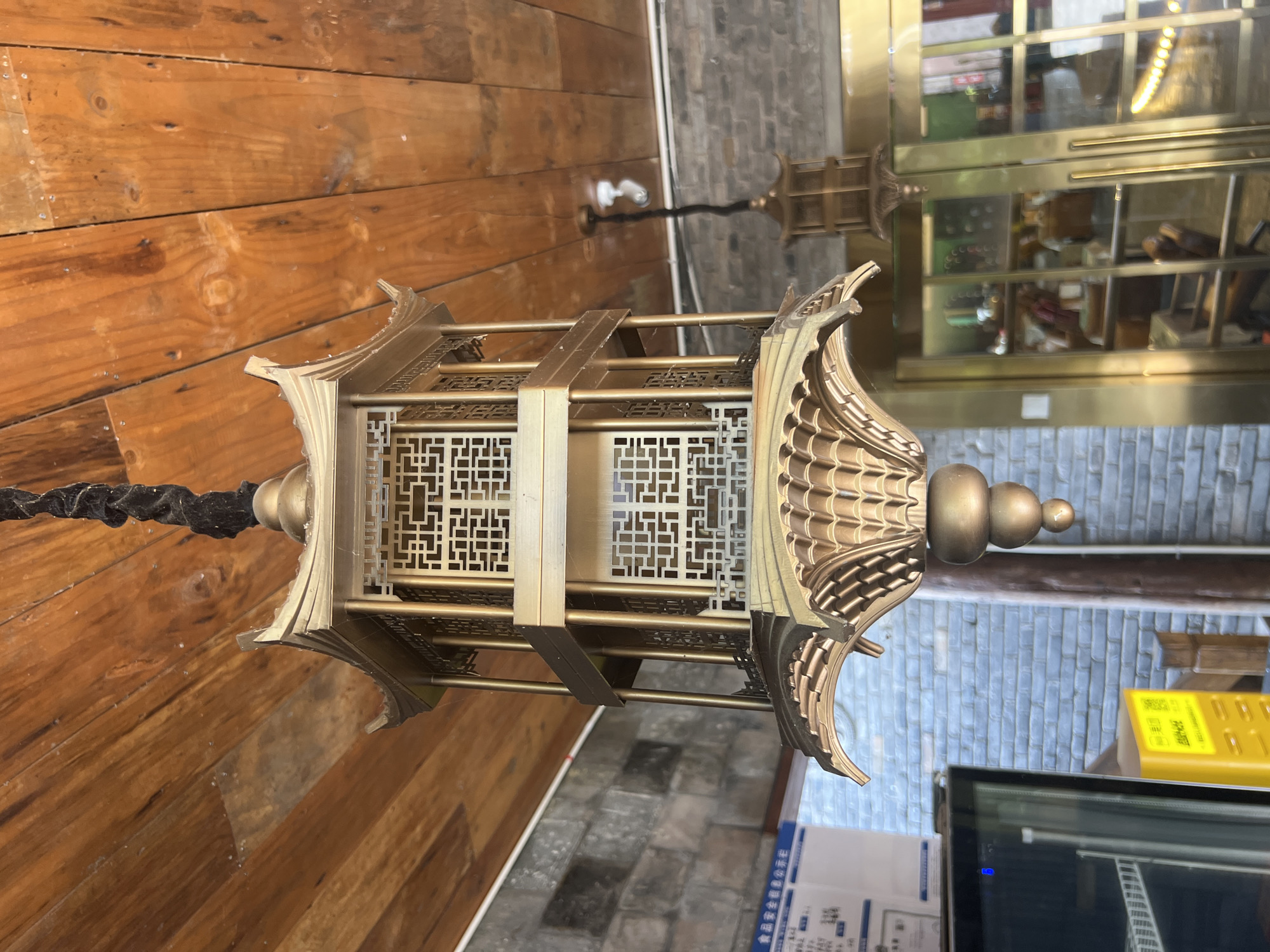 A multi-tiered, pagoda-style lantern made of gold-colored metal hangs from a wooden ceiling. The lantern has intricate latticework on each tier and is suspended by a thick black cord. In the background, a brick wall and a glass-doored cabinet filled with items are visible. Another smaller, similar lantern hangs further in the background. The scene appears to be indoors, possibly in a restaurant or shop in Yuzhong District, China.