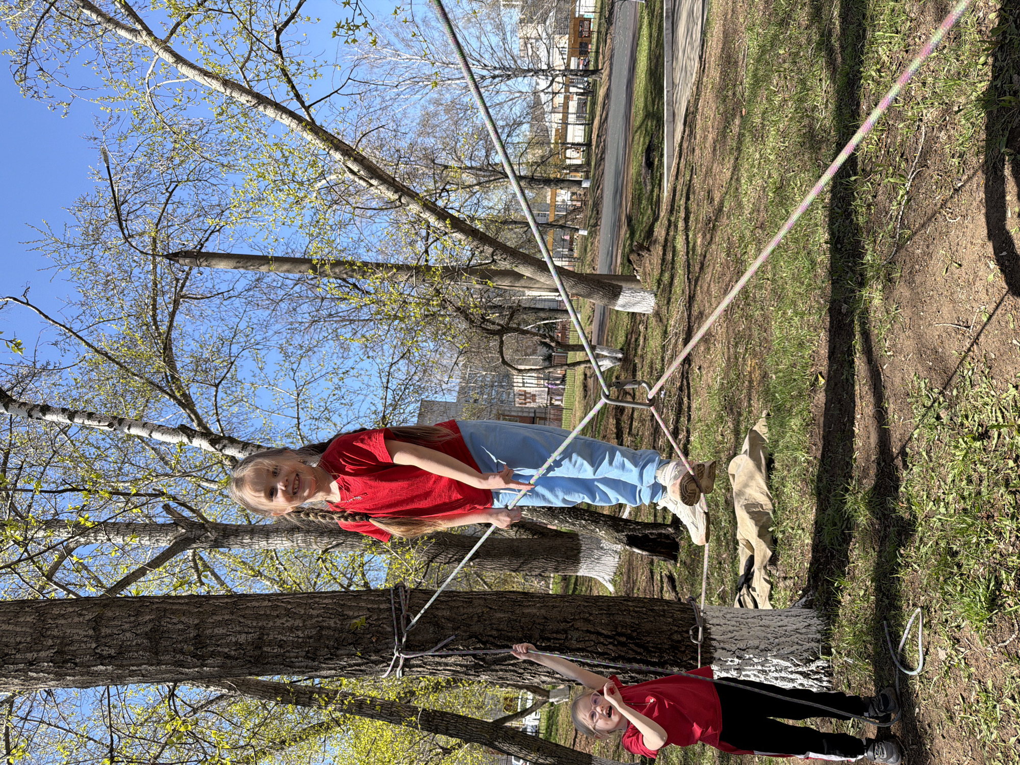 Two girls are playing on a homemade rope climbing structure attached to trees in an outdoor urban area, possibly a park or yard. The scene is in Izhevsk, Russia. The first girl, dressed in a red shirt and blue pants, is standing on the structure. The second girl, wearing a red shirt and black pants, stands on the ground holding a rope connected to the structure. The structure itself is comprised of ropes tied to trees. The ground is covered with grass and dirt, with shadows cast by the trees. In the background, there are residential buildings and a paved street. The sky is blue and sunny.