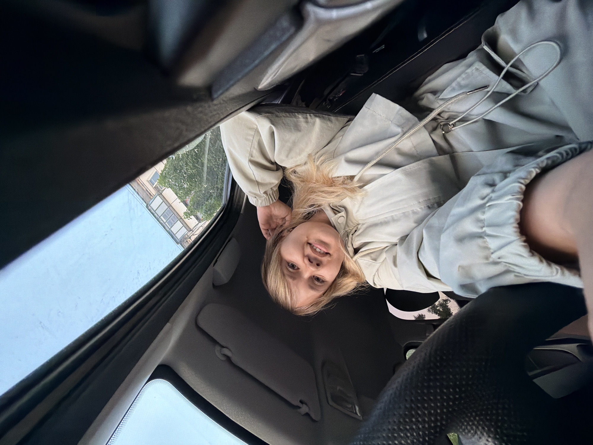 A person with blonde hair, wearing a light-colored jacket, is inside a car. The image is taken from a low angle, looking upwards. Outside the car window, a building can be seen. The location is Chaikovsky, Russia.
