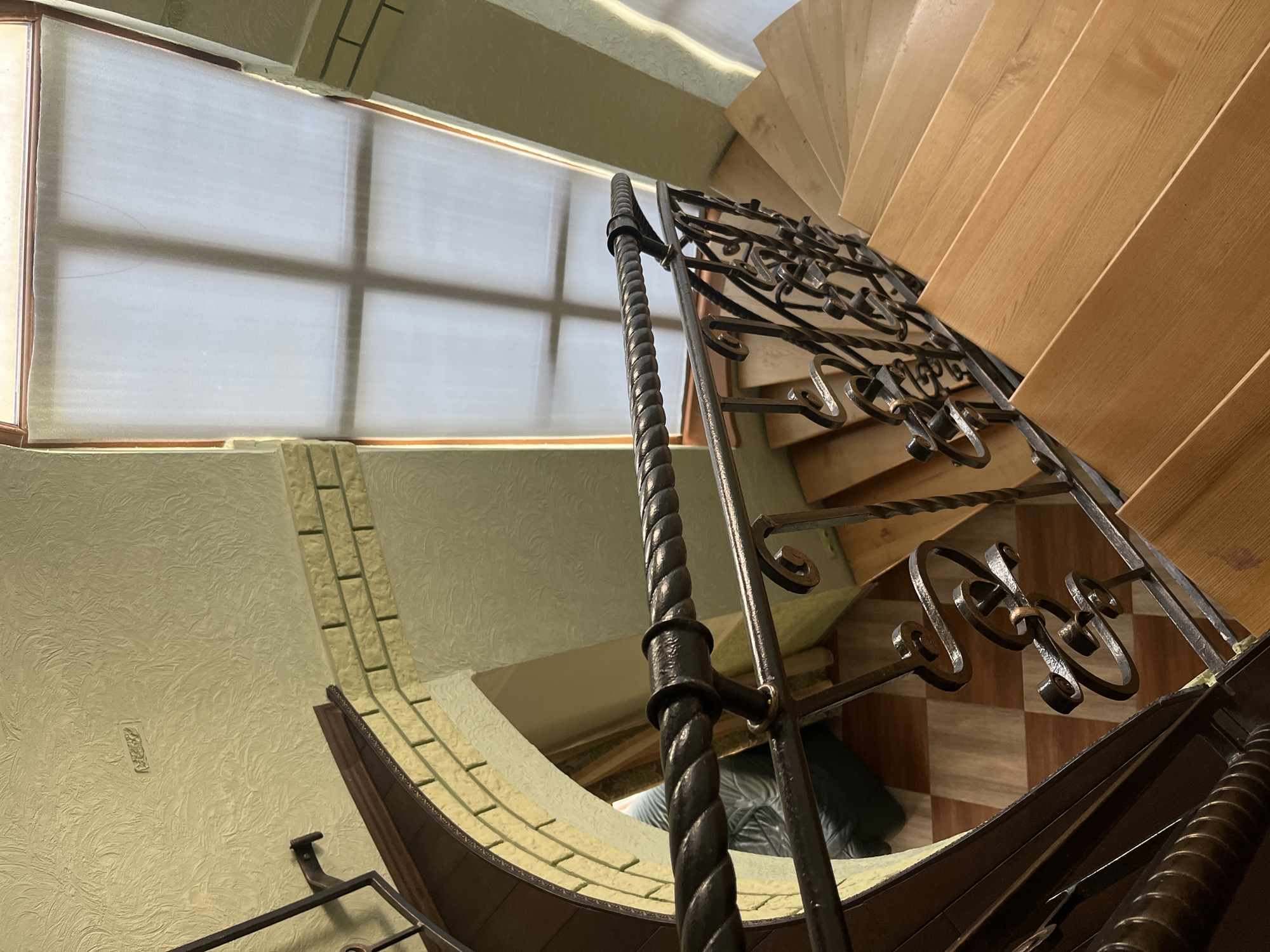 The image depicts a spiral staircase with wooden steps and a black wrought-iron railing. The railing features a thick, twisted handrail and decorative scrollwork. To the left of the staircase, there is a textured yellow wall with what appears to be a decorative brick accent. Above the staircase, a large skylight with a translucent covering diffuses the sunlight. On the landing visible at the bottom of the staircase, the floor is tiled in a checkered pattern of brown and beige squares. The environment appears to be indoors, possibly in a residential building. The scene is static and does not show any people or ongoing actions. The overall lighting suggests daytime. No specific location in Tarnivtsi, Ukraine can be identified from the visual information alone.