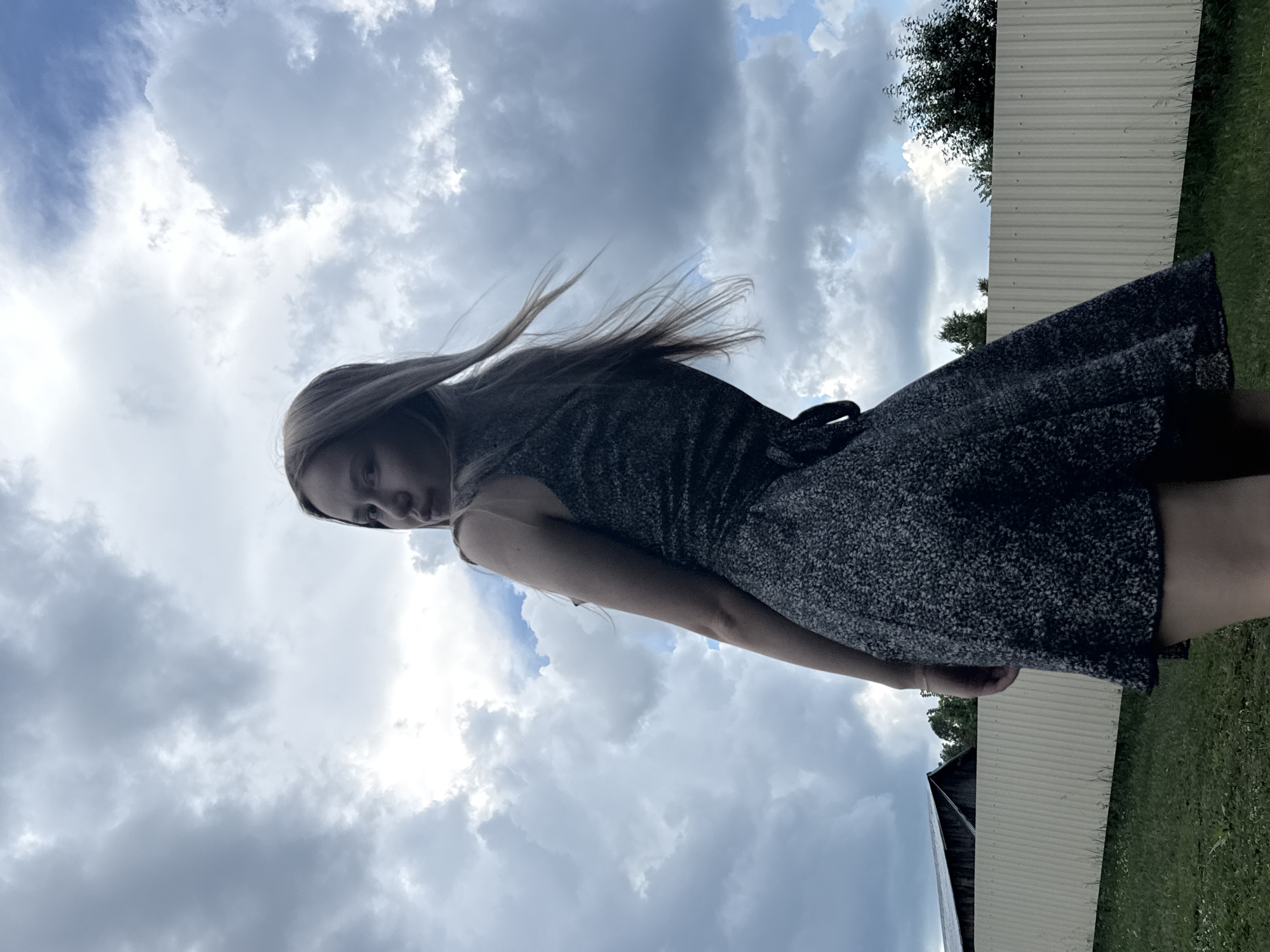 A young woman with blonde hair, wearing a dark, patterned dress, stands outdoors against a cloudy sky. Her body is angled away from the camera, with her head turned to look back over her shoulder. The background includes a light-colored, corrugated metal fence, green grass, and some trees. In the distance, there appears to be a building with a dark roof. The scene is likely in a rural or semi-rural area, possibly on the outskirts of Zhigalki, Russia, given the presence of natural elements and agricultural-style fencing. The lighting suggests it is daytime, with the clouds creating a diffused light.