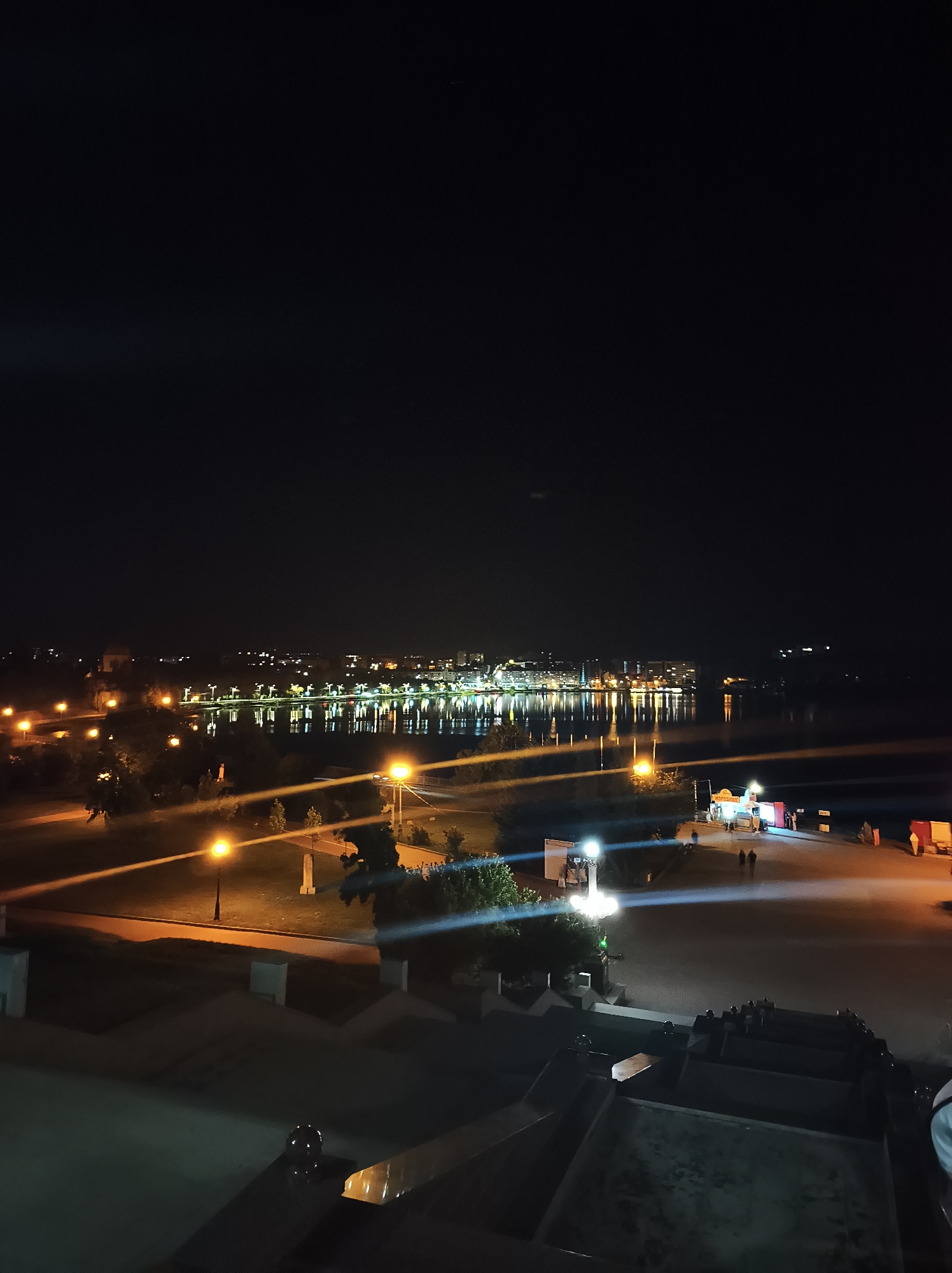 The image shows a nighttime view of the Ternopil pond and the surrounding area in Ternopil, Ukraine. The pond reflects the lights of the city, creating a shimmering effect. Several streetlights illuminate the pathways and green spaces along the water's edge. In the distance, the lights of buildings are visible, suggesting an urban environment. A few people can be seen walking along the promenade. The foreground shows a raised area with some architectural features, possibly a balcony or observation deck, from which the photograph was taken. The overall scene conveys a peaceful and illuminated atmosphere of the city at night.