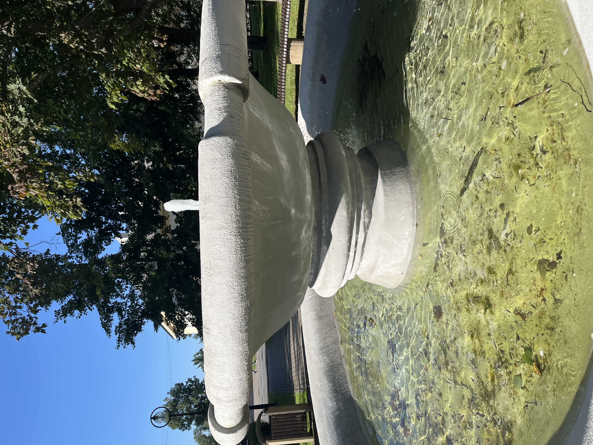 In the city of Kohtla-Järve linn, Estonia, a public fountain stands under a clear blue sky. The fountain is made of concrete and features a tiered design with a wide basin. Water spouts from the center, creating a small spray. The basin of the fountain is filled with water that appears somewhat murky and contains fallen leaves and debris. Behind the fountain, a paved walkway is visible, bordered by green grass and trees. In the distance, a bench and a trash receptacle can be seen, suggesting a park or public area. The overall scene depicts a quiet, outdoor environment on a sunny day.