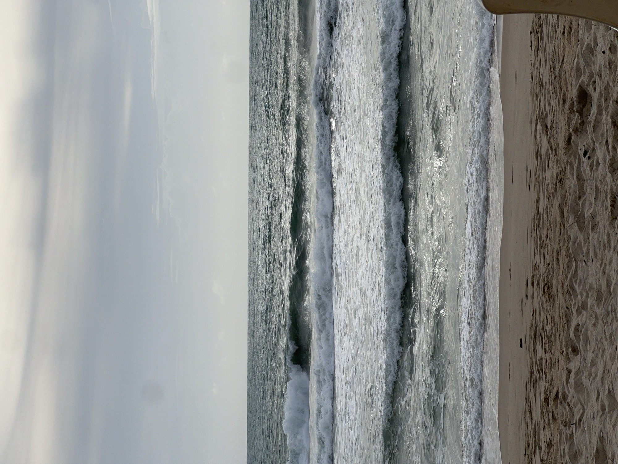 The image depicts a beach scene with the ocean in the background and sand in the foreground. Multiple waves are breaking on the shore, creating white foam. The sky is overcast with clouds. The location is identified as the city of Choeng Thale, Thailand. There are no people or significant objects visible in the frame, except for a sliver of what appears to be a beach chair on the far right edge of the image.