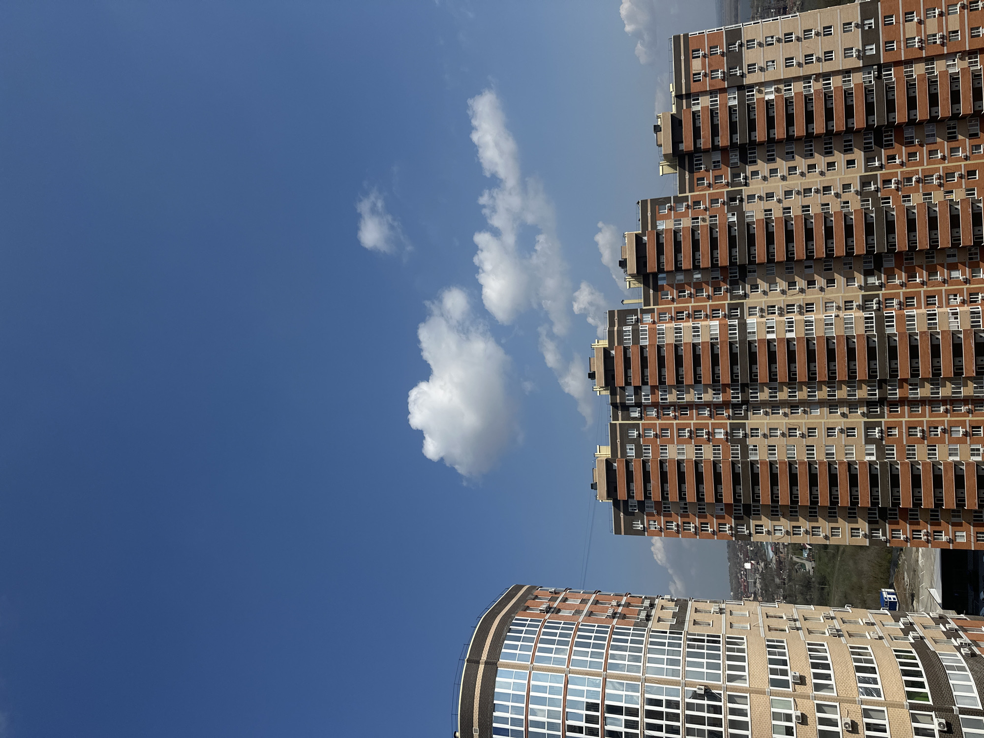 The image shows a view of buildings in Novoaleksandrovka, Russia, against a bright blue sky with some white clouds. There are two main building structures visible: a tall, rectangular stepped building with many windows and air conditioning units, and a curved building with a similar window pattern. The rectangular building features a brick and beige color scheme, while the curved building is primarily beige. In the distance, a glimpse of the city landscape can be seen between the two buildings.