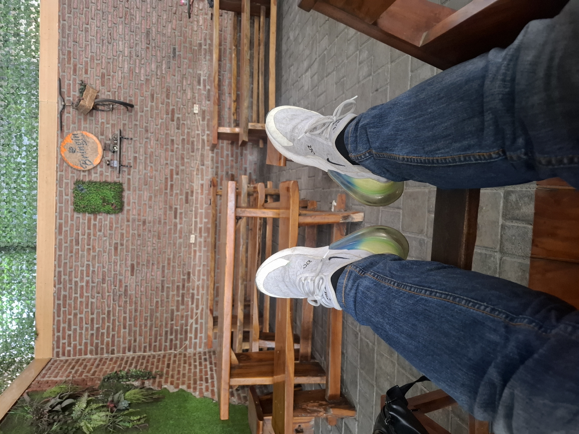 The image depicts a person's legs and feet, clad in blue denim jeans and grey athletic shoes, resting on a wooden chair. The shoes feature a distinctive translucent sole with a gradient of green and yellow colors. In the background, there are several wooden picnic tables and benches arranged on a paved surface. A brick wall forms the backdrop, adorned with greenery, a circular sign reading "Bringin' Inn," a small shelf with two decorative items, and a bicycle with a basket. The scene appears to be an outdoor seating area of a cafe or restaurant, possibly located in Lamongan, Indonesia, given the presence of the "Bringin' Inn" signage. The lighting suggests it is daytime.