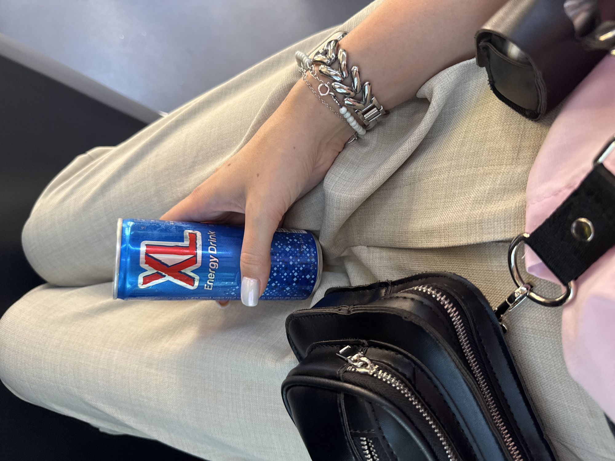 A person's right hand, with white nail polish and silver bracelets, is holding a blue can of XL Energy Drink. The can has red lettering and a pattern of small white dots. The person is wearing light-colored linen pants. A black purse with zippers is visible on the left side of the image, and a pink item is underneath it. The scene appears to be indoors, possibly in a public waiting area, with dark gray seating. The lighting is natural and soft. No specific location in Holon, Israel can be identified from the visual information provided.