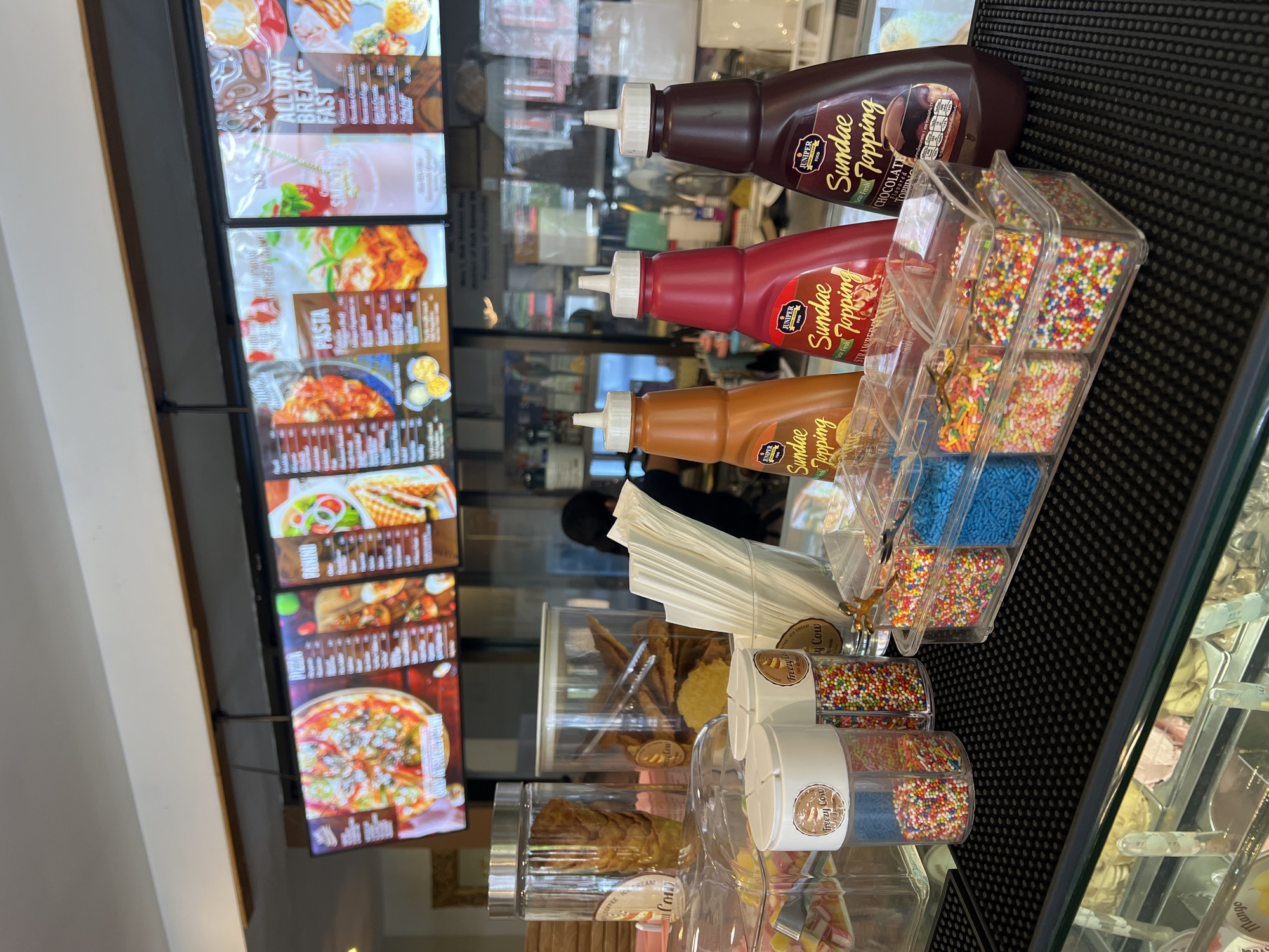 The image shows a counter in a food establishment. Three bottles of Juniper Sundae Topping are displayed on a black surface, flavored chocolate, strawberry, and caramel. Next to them is a clear plastic container divided into sections, each holding different colored sprinkles. Below, there are cylindrical containers with more sprinkles. Paper napkins are stacked in a white container. Behind these items is a reflective glass surface, which reveals a glimpse of a menu board displaying various food items, including breakfast, pasta, and pizza. The logo "Freezy Cow" is visible on the containers. Based on the text, the store's location is in the city of Baan Bo Phut, Thailand.