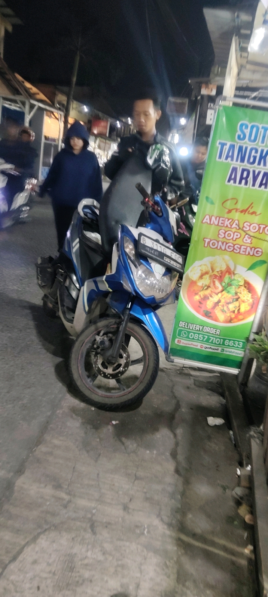 The image depicts a street scene at night in Mekarmukti, Indonesia. In the foreground, a blue and white motorcycle is parked on a cracked asphalt surface. The motorcycle has a license plate that reads "B 3110 FOG". To the right of the motorcycle is a vertical green banner advertising "Soto Tangkar Arya" and other dishes like "Aneka Soto Sop & Tongseng". Below the food offerings, there is a section for "DELIVERY ORDER" with a phone number "0857 7101 6633" and logos for ShopeeFood, GoFood, and GrabFood.
Behind the motorcycle, three individuals are visible. The person on the far left is partially obscured by the motorcycle. In the center, a person wearing a dark blue hooded jacket stands looking forward. To their right, a person in a dark jacket with a green logo on the chest is also looking forward. The background shows a dimly lit street with buildings and possibly more vehicles. The overall lighting suggests it is nighttime, with artificial lights illuminating the scene.