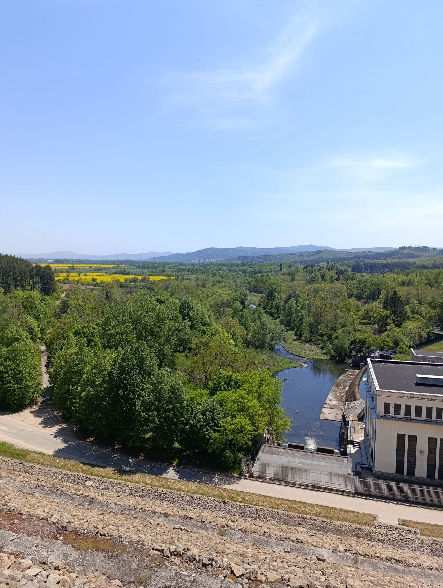 The image presents a high-angle view of a landscape in an unspecified city. The foreground consists of a dam structure built from rocks and concrete. Adjacent to the dam is a paved road that curves around a cluster of trees.
Beyond the road, the view opens up to a river surrounded by dense trees. An industrial building with a flat roof and rows of windows sits next to the river, possibly part of a hydroelectric facility. In the background, there are fields, some of which appear to be filled with yellow flowers, and distant mountains under a clear blue sky with light clouds.