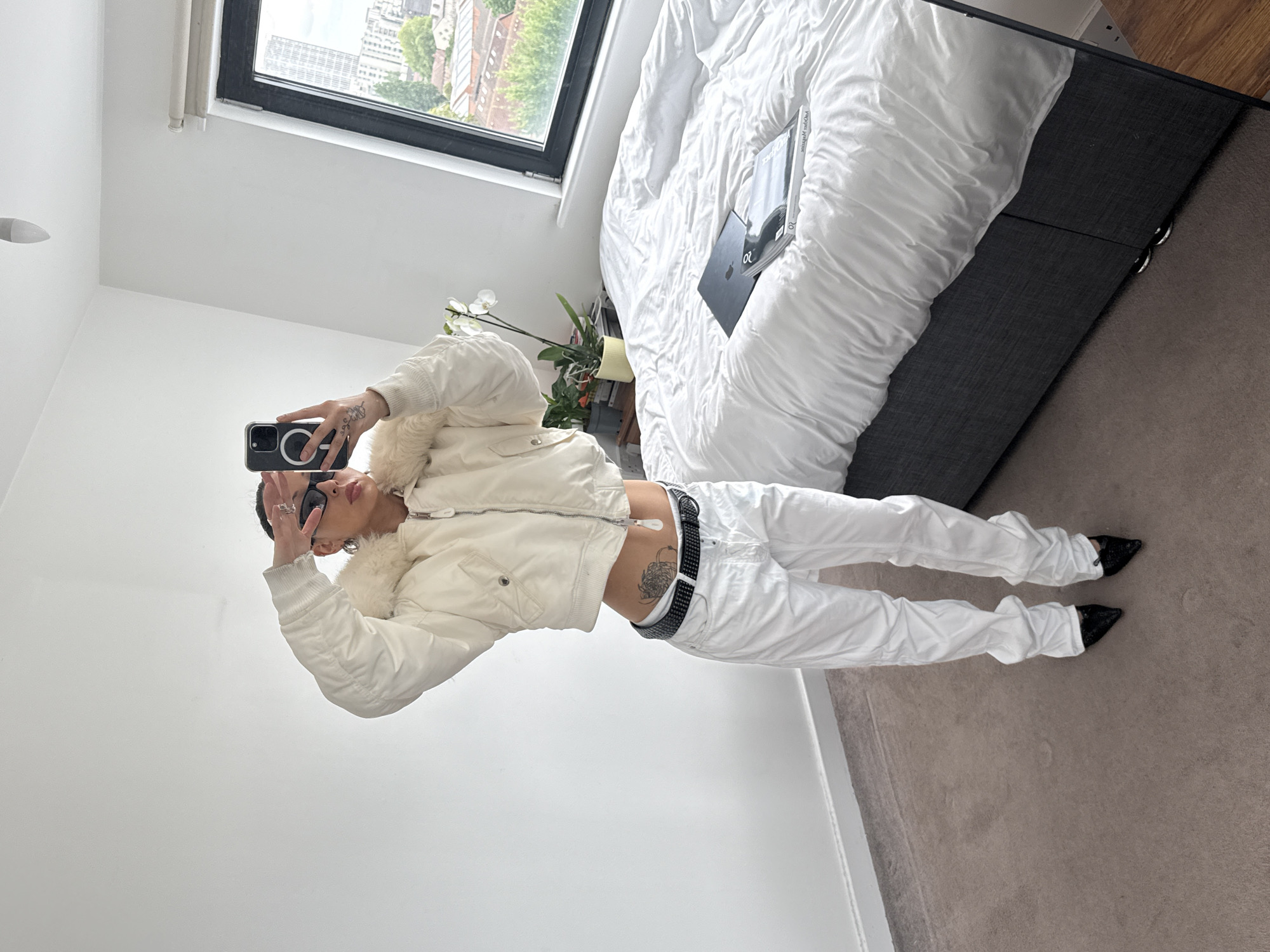 The image depicts a person taking a selfie in a modern, brightly lit room. The person is wearing a white jacket with fur trim, white pants, and black shoes, and they are holding a smartphone. The background includes a window with a view of a city landscape, a bed with white linens and a book, and a plant on a table. Based on the architecture and greenery visible through the window, the location is likely in London, United Kingdom.