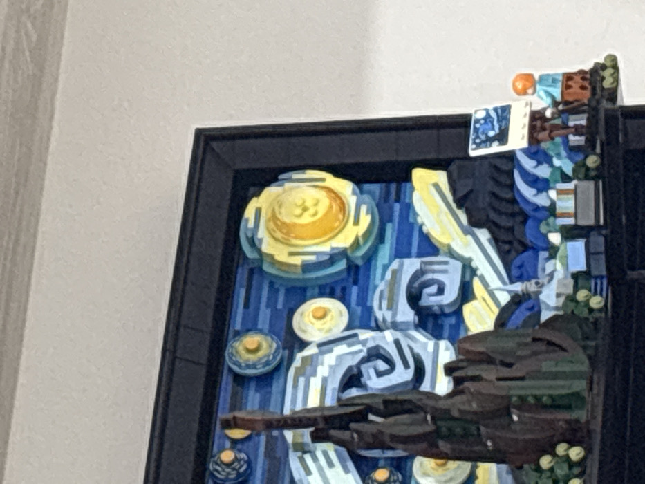 A framed artwork, seemingly a recreation of Van Gogh's "The Starry Night," is predominantly blue and yellow. It features a large swirling yellow sun, smaller celestial bodies, and swirling clouds. A dark brown, tree-like structure is in the lower right. The artwork appears to be constructed from small, possibly brick-like components. The frame is black. The background is a plain white wall.