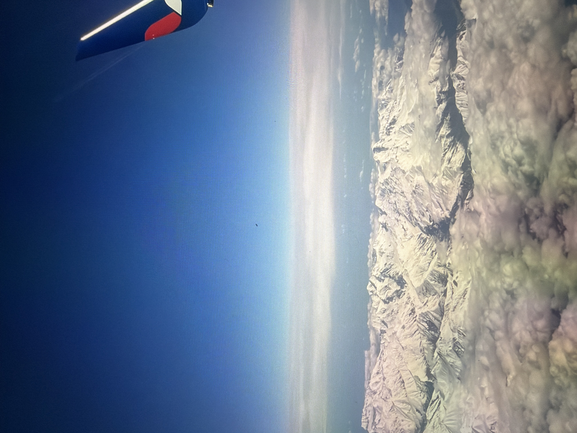 An aerial view captures snow-capped mountains beneath a clear blue sky. An airplane tail fin, predominantly blue with a red and white emblem, is visible in the upper right corner of the frame, indicating the perspective is from an aircraft in flight. The lower half of the image shows a vast expanse of jagged, snow-covered mountain peaks partially obscured by fluffy white clouds. The light suggests it is daytime, with the sun illuminating the scene from above. The image does not contain any elements that would suggest a specific location in the city of Kotelniki, Russia, as it depicts a natural landscape from a high altitude.