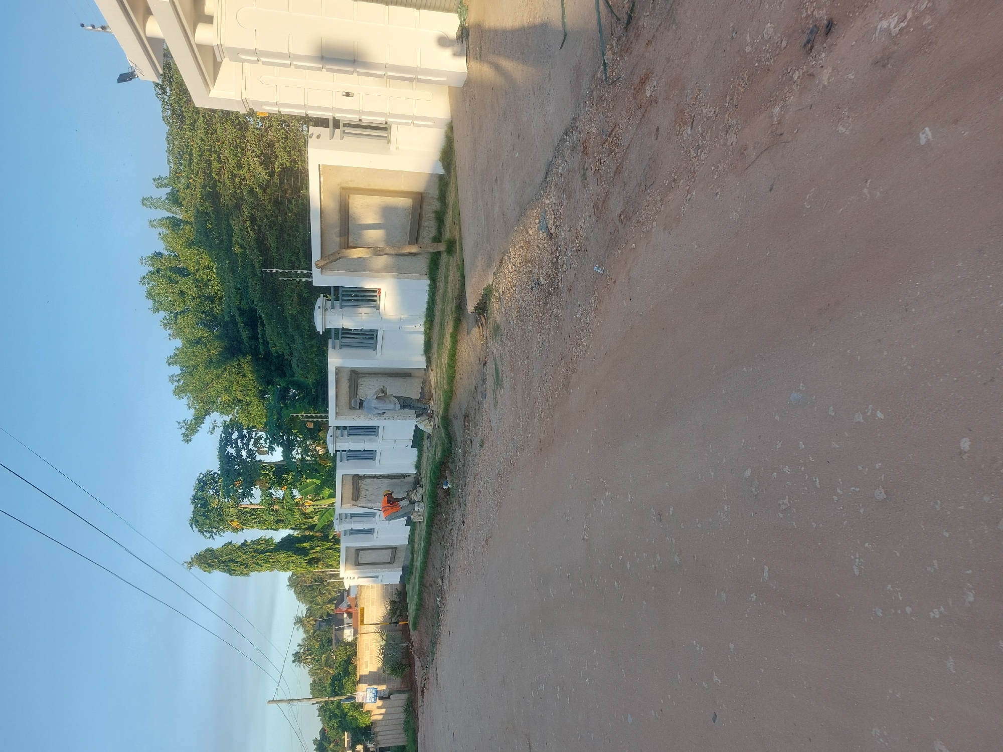 The image depicts a dusty, unpaved road in a residential area of Dar es-Salaam, Tanzania. On the left side of the road are several white, multi-story buildings in various states of completion, some appearing newly constructed. Green foliage, including trees and smaller plants, is present between the buildings. Two individuals can be seen near the buildings, one wearing an orange shirt and another a gray shirt. They appear to be engaged in construction or maintenance work. The sky is clear and blue. Utility poles and wires are visible on the left side. In the background, other buildings and greenery can be observed. The road occupies a significant portion of the foreground and appears well-used.