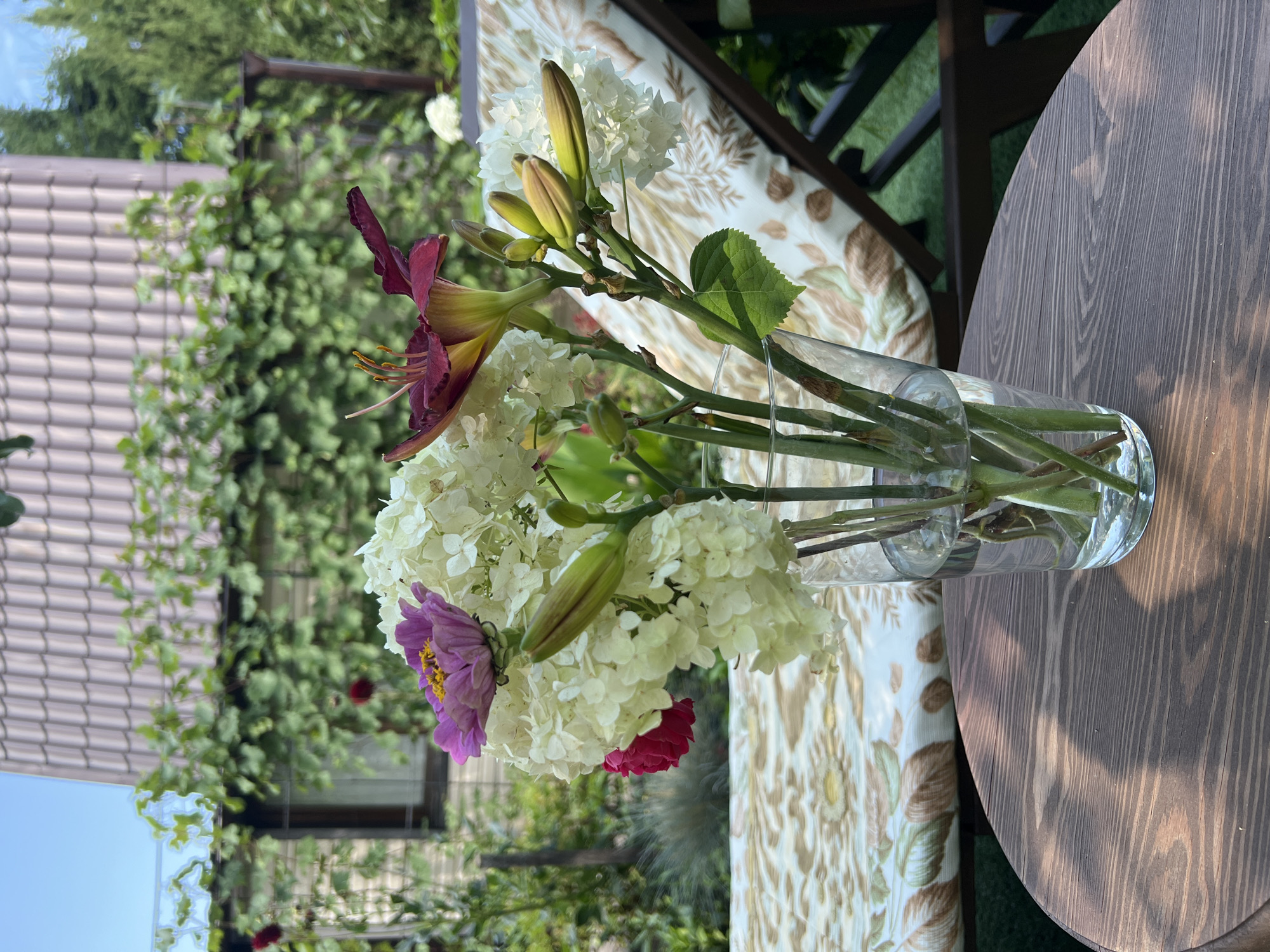 A close-up, eye-level shot captures a bouquet of flowers in a glass vase, placed on a circular wooden table. The bouquet features white hydrangeas, a pink zinnia, and a dark red daylily. Behind the table, a cushioned lounge chair with a floral pattern is visible. In the background, a tiled roof of a house and green foliage suggest a garden setting. The lighting indicates it is daytime. The image does not contain any people or indicate any specific actions or interactions. The location is Lopatino, Russia.