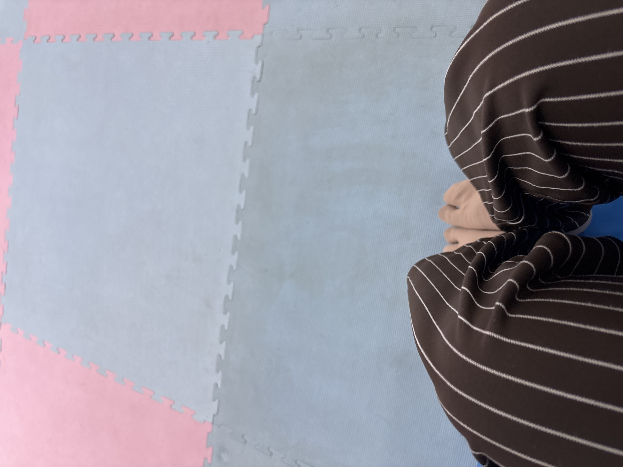 This is a first-person perspective shot of someone's legs and hands resting on a blue mat. The person is wearing brown pants with white pinstripes. The mat is part of a larger interlocking mat surface, with sections of light grey and pink visible in the background. The overall environment appears to be an indoor recreational or training space. The specific location in the city of Liangjulang, Indonesia, cannot be determined from the image alone.