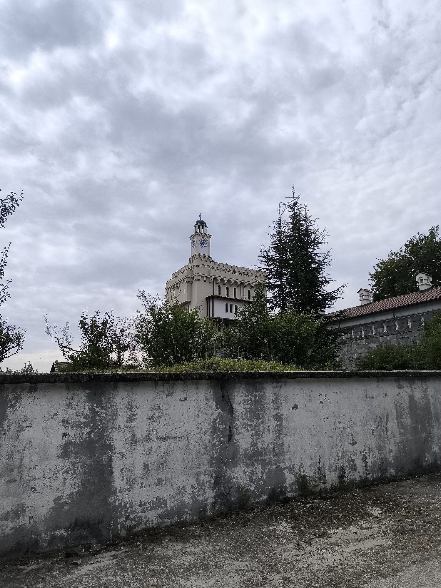 A beige fortress-like building with a clock tower stands behind a stone wall and trees. The sky is overcast with gray clouds. The wall in the foreground is weathered and textured, with patches of peeling paint. A gravel path is visible in front of the wall. The building appears to be in the city of Hadzhidimovo, Bulgaria, as suggested by external information. No people or animals are visible in the image.