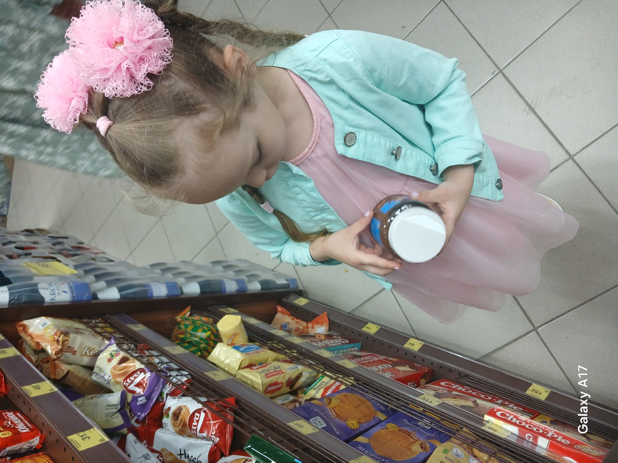 Here is a detailed description of the image:
A young girl wearing a light blue jacket and a pink dress is looking down at a jar she is holding. The jar has a brown label and a white lid. She has blonde hair with two pink flower hair clips. The scene appears to be inside a store aisle, possibly in Yekaterinburg, Russia, given the Cyrillic text on the product labels. The aisle is filled with various snack products, including cookies and cakes. The products are arranged on shelves with price tags and product names visible. Some brands that can be identified include "Days" cookies and "Choco-Pie." The floor is tiled. A portion of a shopping cart or another person is visible on the left side of the frame.