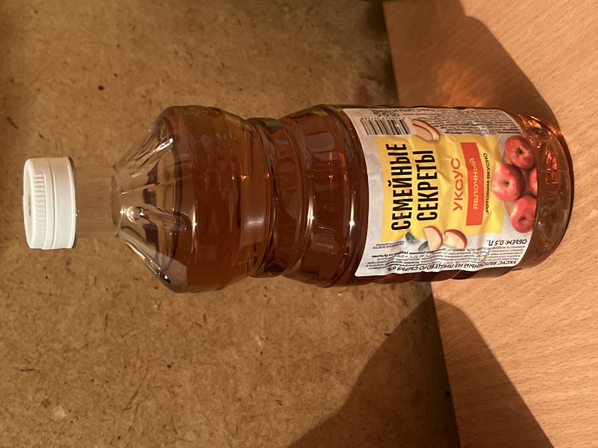 A clear plastic bottle of apple cider vinegar, labeled in Russian, is placed on a wooden surface. The label features yellow and red text and illustrations of apples. The bottle has a white screw-on cap. The background is a textured brown surface. The scene appears to be indoors, possibly on a table or shelf. There is no indication of the city of Iksha, Russia, in the image.