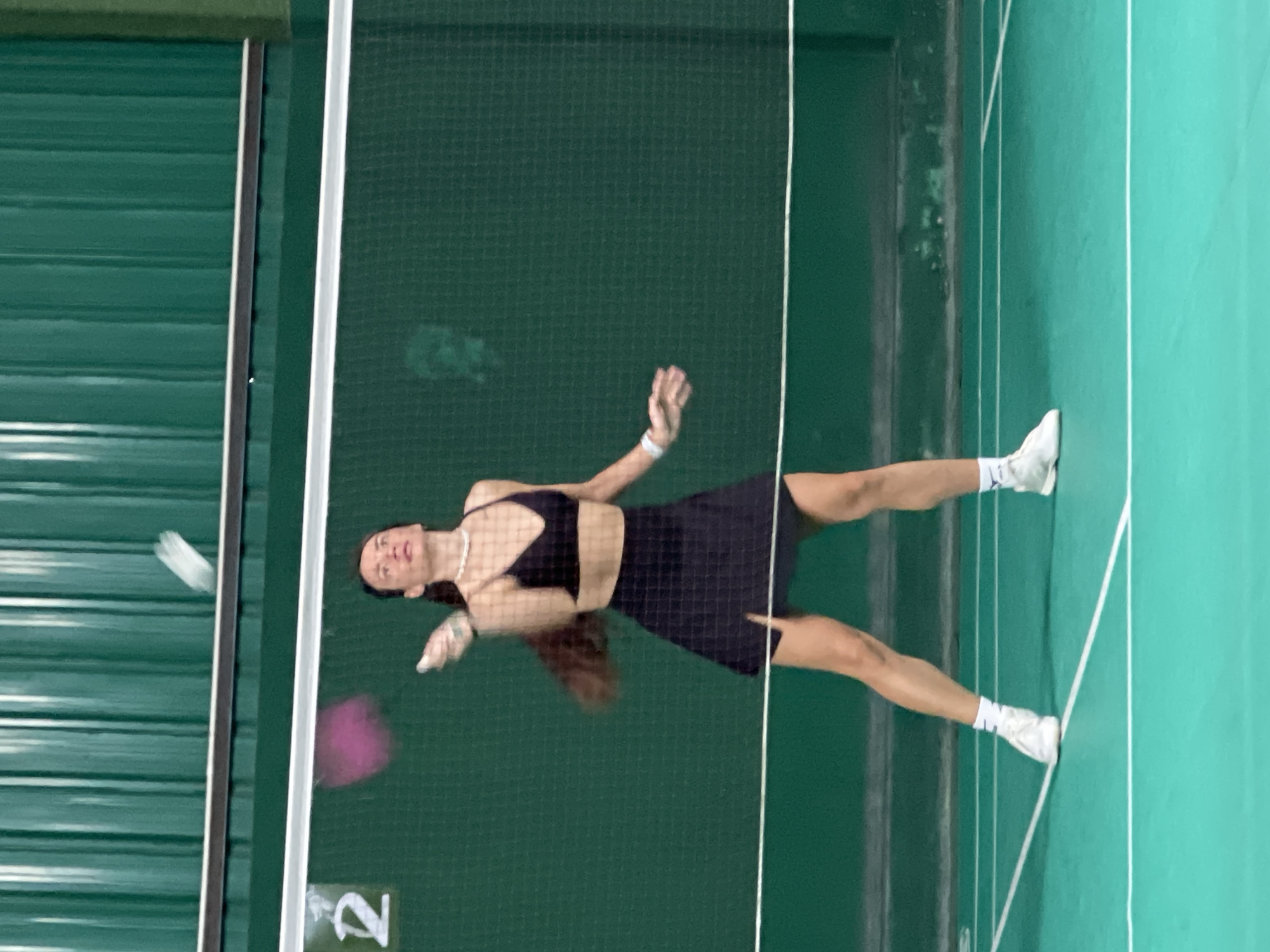 A woman in a black sports bra and skirt is playing badminton indoors. She is wearing white athletic shoes and a pearl necklace. A badminton shuttlecock is in the air near the net. The location is a badminton court, possibly in or near บ้านบางรักษ์, Thailand.