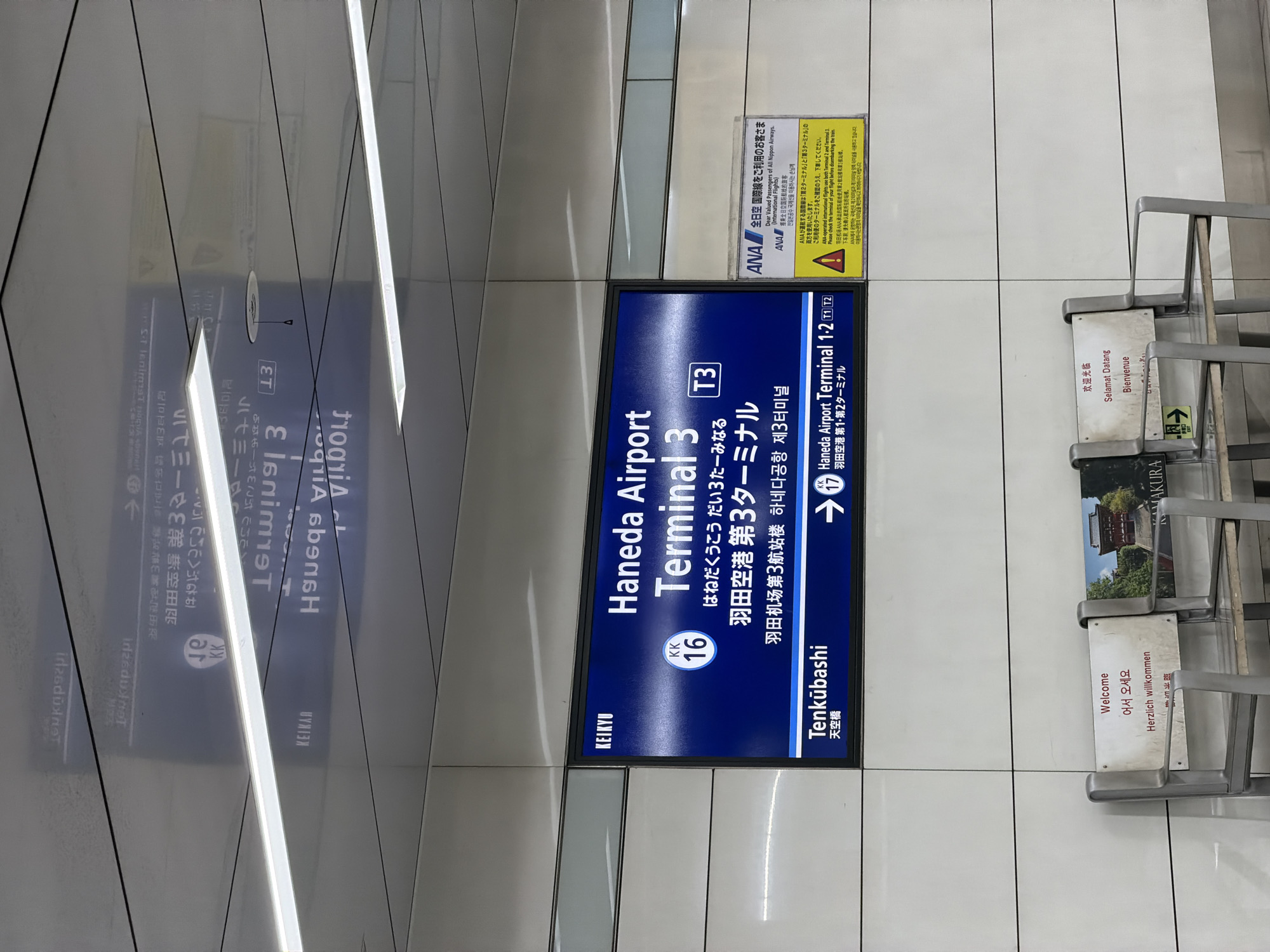 The image shows a subway or train station interior with signage and seating. A large blue sign displays information about "Haneda Airport Terminal 3" in English, Japanese, and Korean, with a "KEIKYU" logo at the top left. The sign indicates connections to "Tenkubashi" station, and also mentions "Haneda Airport Terminal 1.2". To the right of the blue sign is a yellow information placard from ANA (All Nippon Airways) regarding international flights to terminals 2 and 3. In the foreground, there are two metallic benches with signs on them displaying "Welcome" in multiple languages including English, Korean, and German. The environment appears to be modern and clean, with white tiled walls and illuminated ceiling panels. The lighting is bright, likely from overhead fixtures. The scene depicts an indoor transportation hub, likely within Ota, Japan, given the explicit mention of Haneda Airport. There are no people visible in the image.