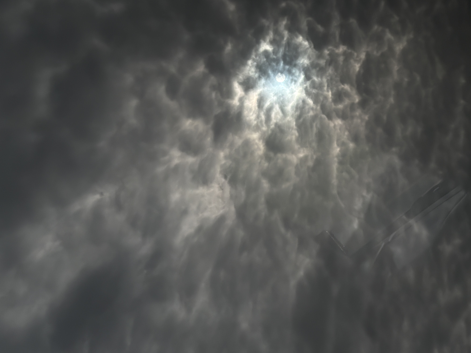 The image depicts a cloudy sky with the sun partially obscured by the clouds. The clouds are dark gray and textured. In the lower right, there's a faint, translucent form resembling an airplane. There is no information to describe the location in the city of Jouy-le-Moutier, France. There are no people, specific subjects, objects, actions, or interactions discernible in this image, only the sky and clouds.