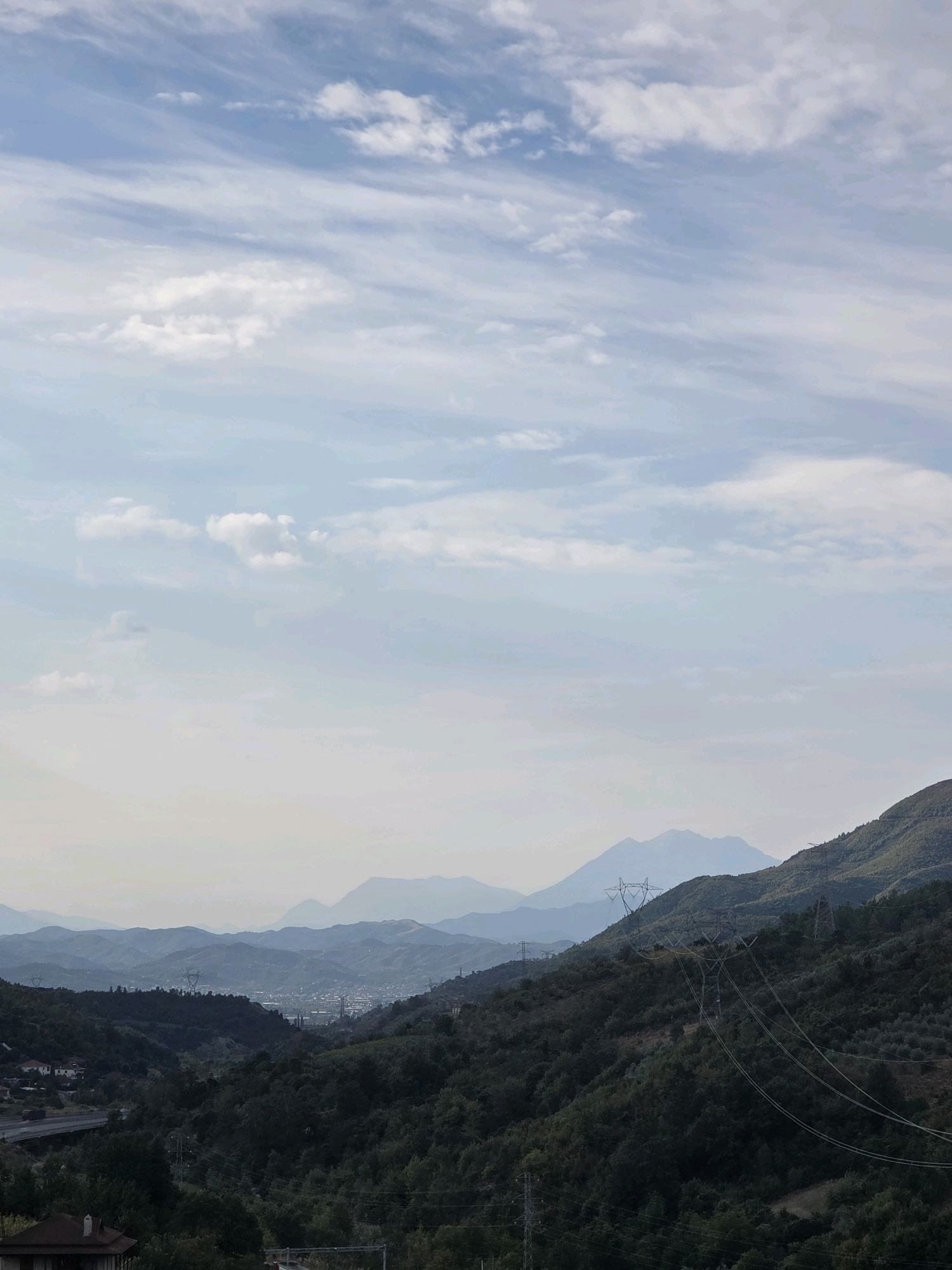 The image captures a panoramic view of the landscape surrounding Gracen, Albania. The sky is filled with wispy clouds against a light blue backdrop. In the foreground, dense green trees cover rolling hills, with power lines and transmission towers intermittently visible on the slopes. A road and some houses are nestled on the left side of the frame. Further into the distance, a city or town is visible in the valley, with buildings and more industrial-looking structures. Beyond the town, several layers of mountains fade into a hazy, bluish distance, creating a sense of depth and scale. The overall scene depicts a transition from a populated area to a more natural, mountainous terrain.