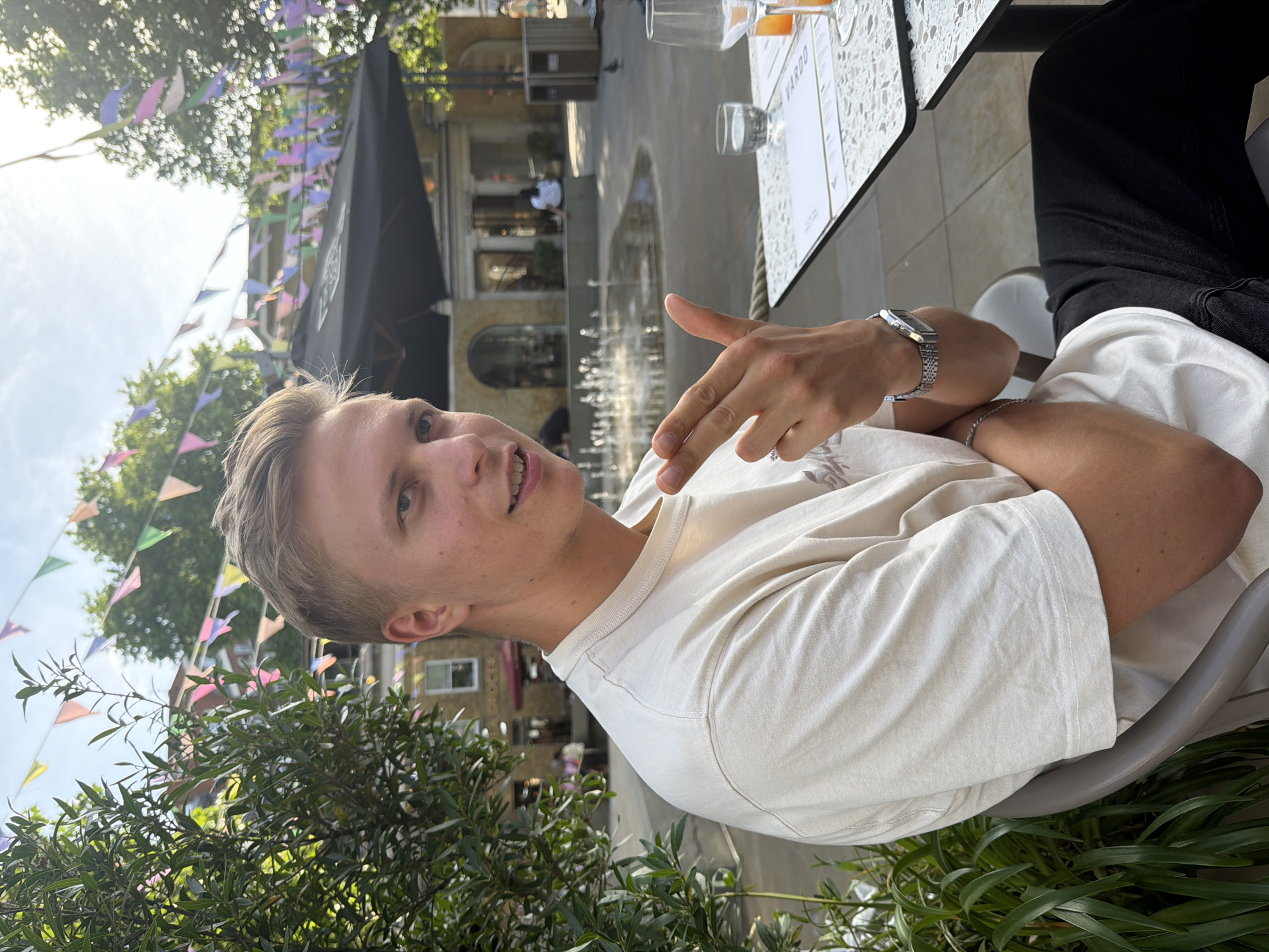 The image captures a young man seated at an outdoor table in what appears to be a public space in London, United Kingdom. He is light-skinned, with blond hair, wearing a light-colored t-shirt and a silver wristwatch on his left wrist. His left arm is bent, with his hand pointing towards the side. He is looking towards the camera with a slight smile.
Behind him, there is a view of a plaza with water fountains and classical-style architecture. Above, there are colorful triangular pennants strung across the area, suggesting a festive or decorative atmosphere. A black awning or structure is visible in the background, possibly part of a building or establishment. The table in front of him contains glasses and a menu. To the side, there are black pants. The image is taken from a slightly low angle. Bushes and plants partially frame the bottom of the shot.