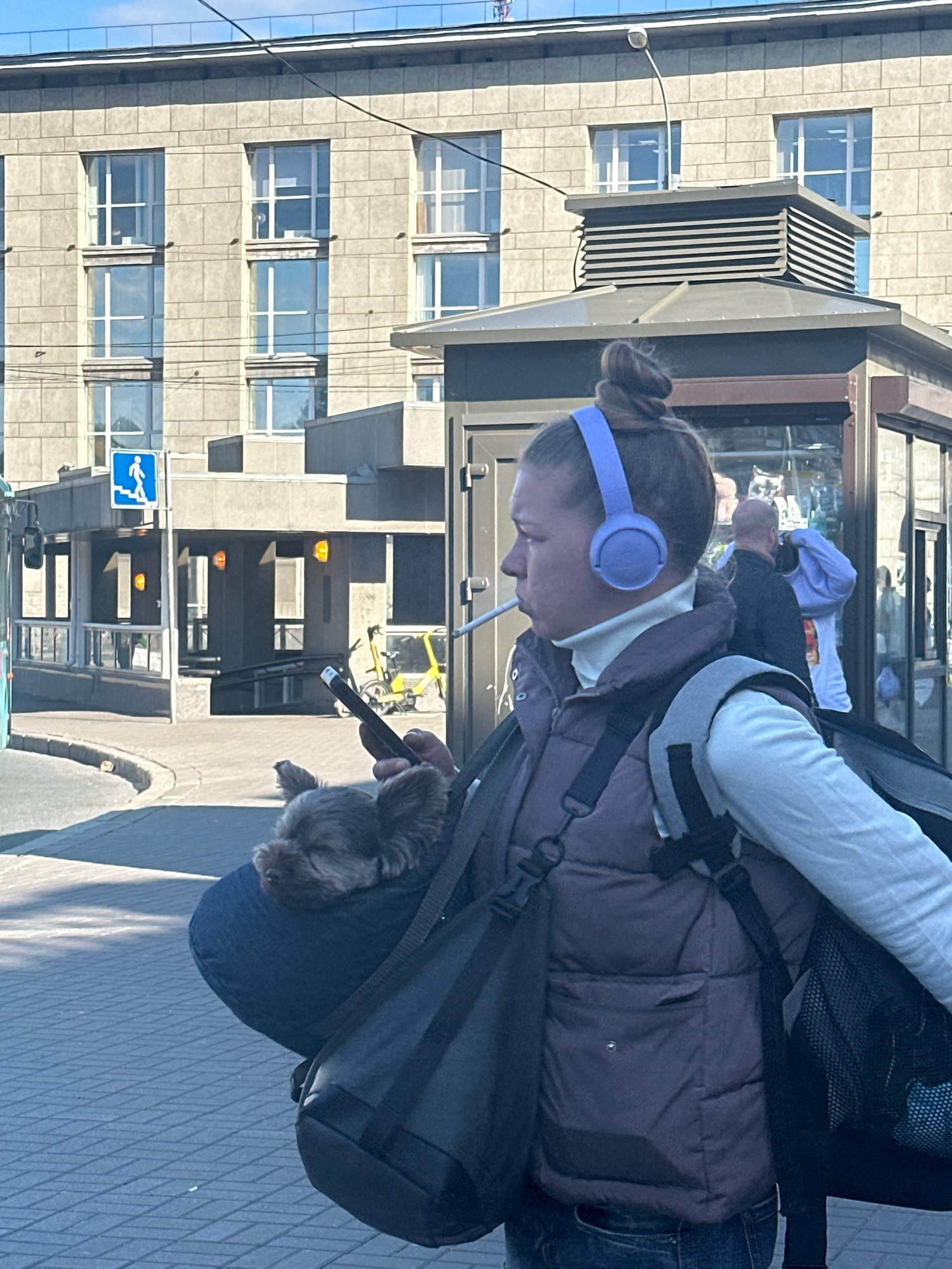 A woman with light brown hair pulled into a bun, wearing a white turtleneck, a brown puffer vest, and jeans, walks outside in Saint Petersburg, Russia. She has light blue headphones on and is holding a cigarette in her mouth. She is also carrying a dark blue bag over her shoulder, with a small, dark-colored dog peeking out of it. The woman is looking to the right, and her right hand is holding a phone. Behind her, a grey building with large windows and a street kiosk are visible. The sidewalk is paved with grey bricks, and a yellow bicycle is parked in the background. The sky is blue and clear.