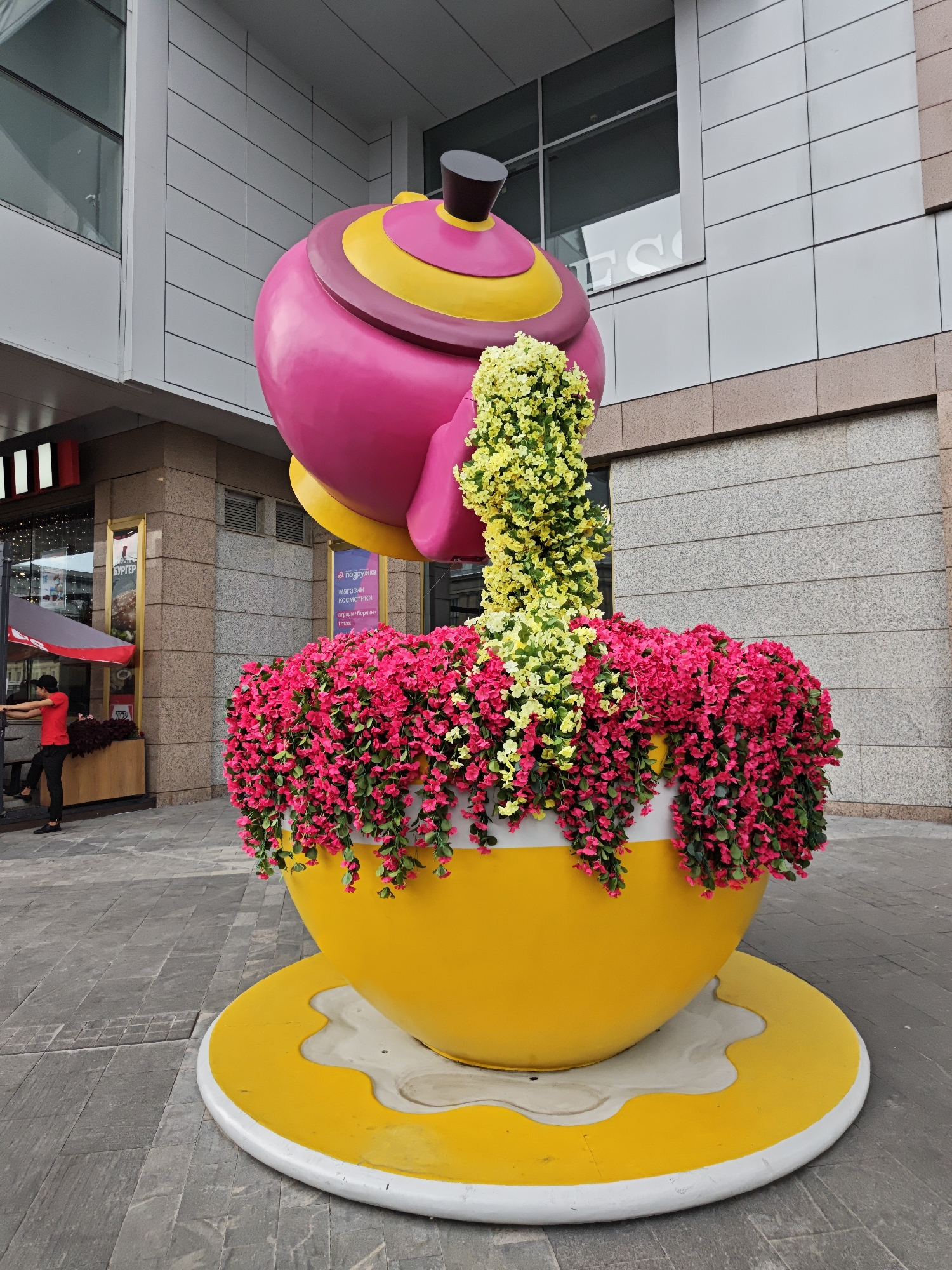 A bright pink and yellow teapot sculpture is overflowing with flowers, appearing to pour them into a large yellow cup and saucer decoration. The teapot has a dark top and is adorned with a yellow band and a darker, possibly burgundy, band. The "tea" flowing from the spout is represented by clusters of bright yellow flowers. The cup is filled with cascading pink flowers, and the saucer is a bright yellow disc with a white, dripped-paint-like design. The entire floral display sits on a grey, paved walkway in front of a modern building with large windows and grey paneled walls. To the left of the sculpture, a person wearing a red shirt and black pants is seen interacting with a counter under an awning. Two signs are visible on the building; one in Russian with text that translates to "Burger" and another that reads "Подружка" (Podruzhka), a cosmetics store. The scene is outdoors in a city environment.