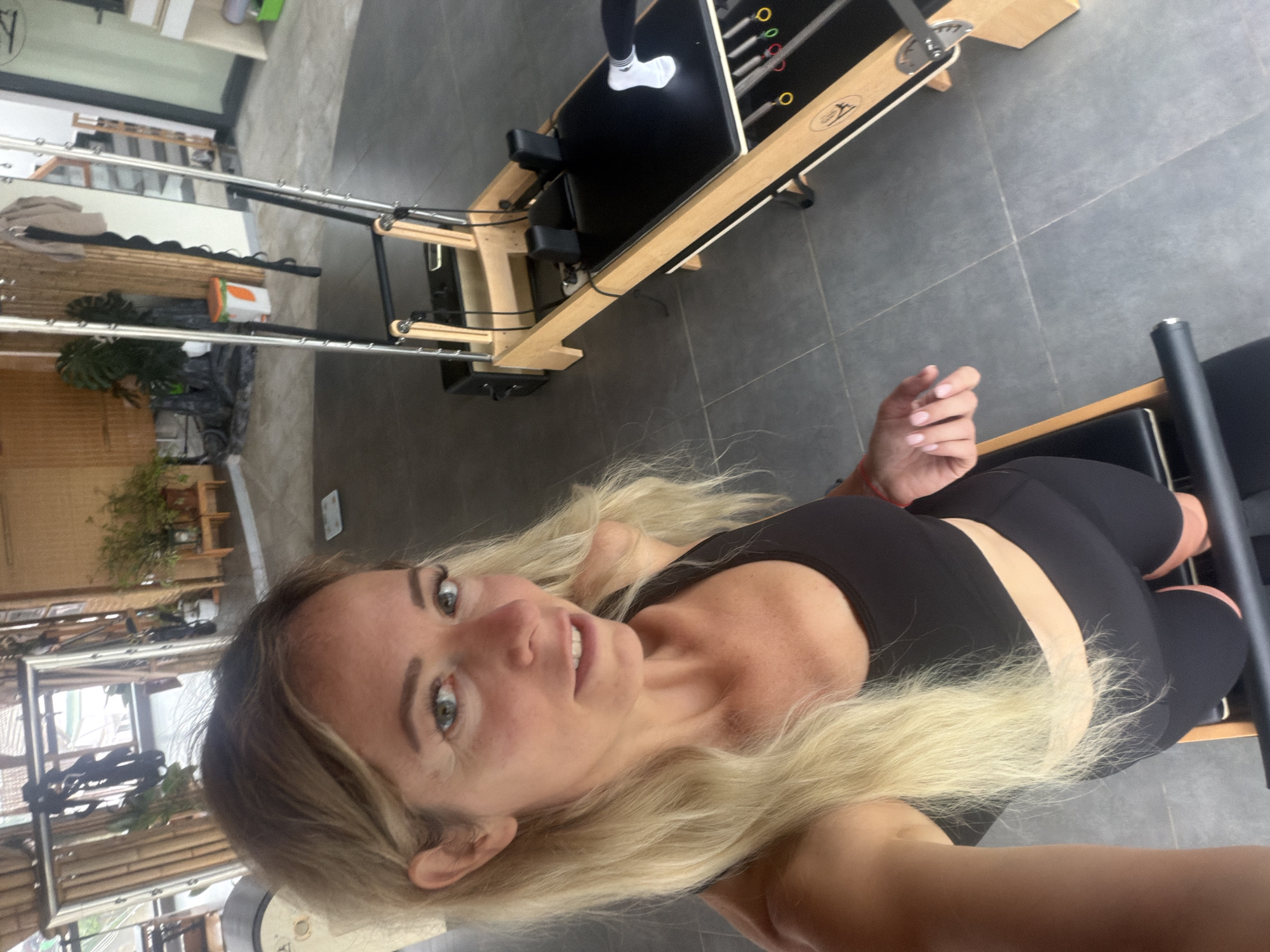 A woman with blonde hair is taking a selfie in a room with a pilates reformer. She is wearing a black sports bra and black shorts. The pilates reformer is made of wood and black upholstery. The room has a tiled floor and bamboo walls. In the background, there are some plants and a mirror.