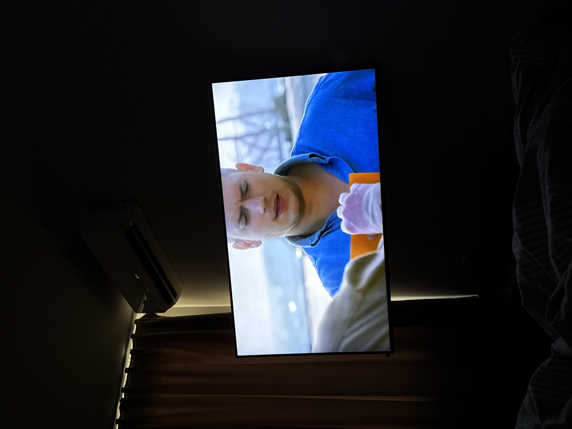 The image captures a dimly lit indoor scene, likely a bedroom or living room, with a large television screen as the focal point. The TV is displaying a close-up of a man with short hair, wearing a blue shirt, whose face shows an expression of distress or pain, with his mouth slightly open. To the right of the man on screen, a hand wearing a light pink or white glove is visible, holding an orange object, suggesting a medical procedure or intense situation within the depicted media.
To the left of the television screen, a white air conditioning unit is mounted on a dark wall, with a small green indicator light illuminated. Below the air conditioner and extending to the left edge of the frame, dark-colored curtains or blinds are partially open, allowing a thin horizontal strip of light to enter from outside, suggesting it might be daytime with the room deliberately darkened. To the right of the television, a dark, rumpled blanket or bedding is partially visible, indicating a bed. The overall lighting of the room is very low, illuminated primarily by the bright glow of the TV screen and the sliver of light from the window. No text is visible in the image. The scene is set in Yasen, Bulgaria.