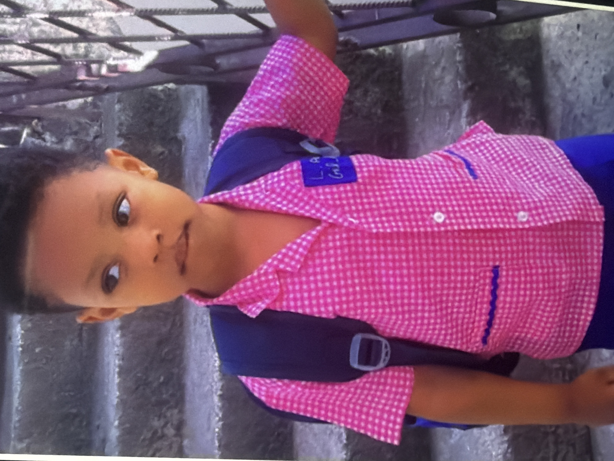 A young child, appearing to be of African descent, stands facing slightly to the right, with their head turned to look at the camera. They are wearing a pink gingham button-down shirt with blue trim on the pockets and a blue backpack. A small blue patch is attached to the left chest of the shirt, with writing that appears to be "L A GRL". The child is standing on grey concrete steps, and a black metal railing is visible to the right. The background shows more concrete steps and a stone wall. The lighting suggests it is daytime. The location is unknown.