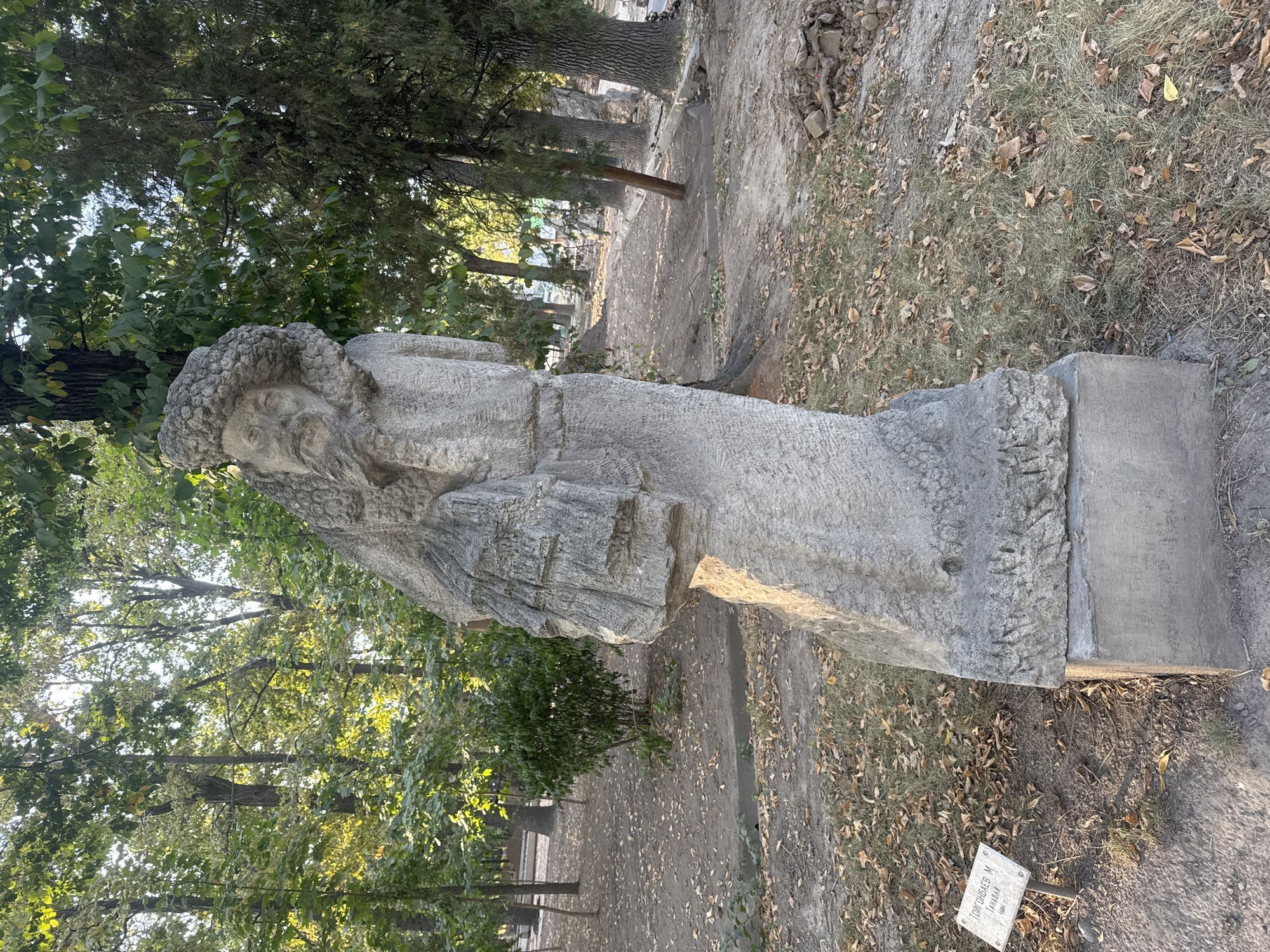 A stone sculpture of a bearded man wearing a fur hat stands in a park in Bishkek, Kyrgyzstan. The sculpture is rough-hewn and appears to depict a historical or mythological figure. The man is holding a book or scroll in his left arm. The sculpture is placed on a concrete base and is surrounded by trees and fallen leaves. A small sign with Cyrillic writing is visible in the lower left corner of the image. The overall scene suggests an outdoor cultural exhibit or memorial.