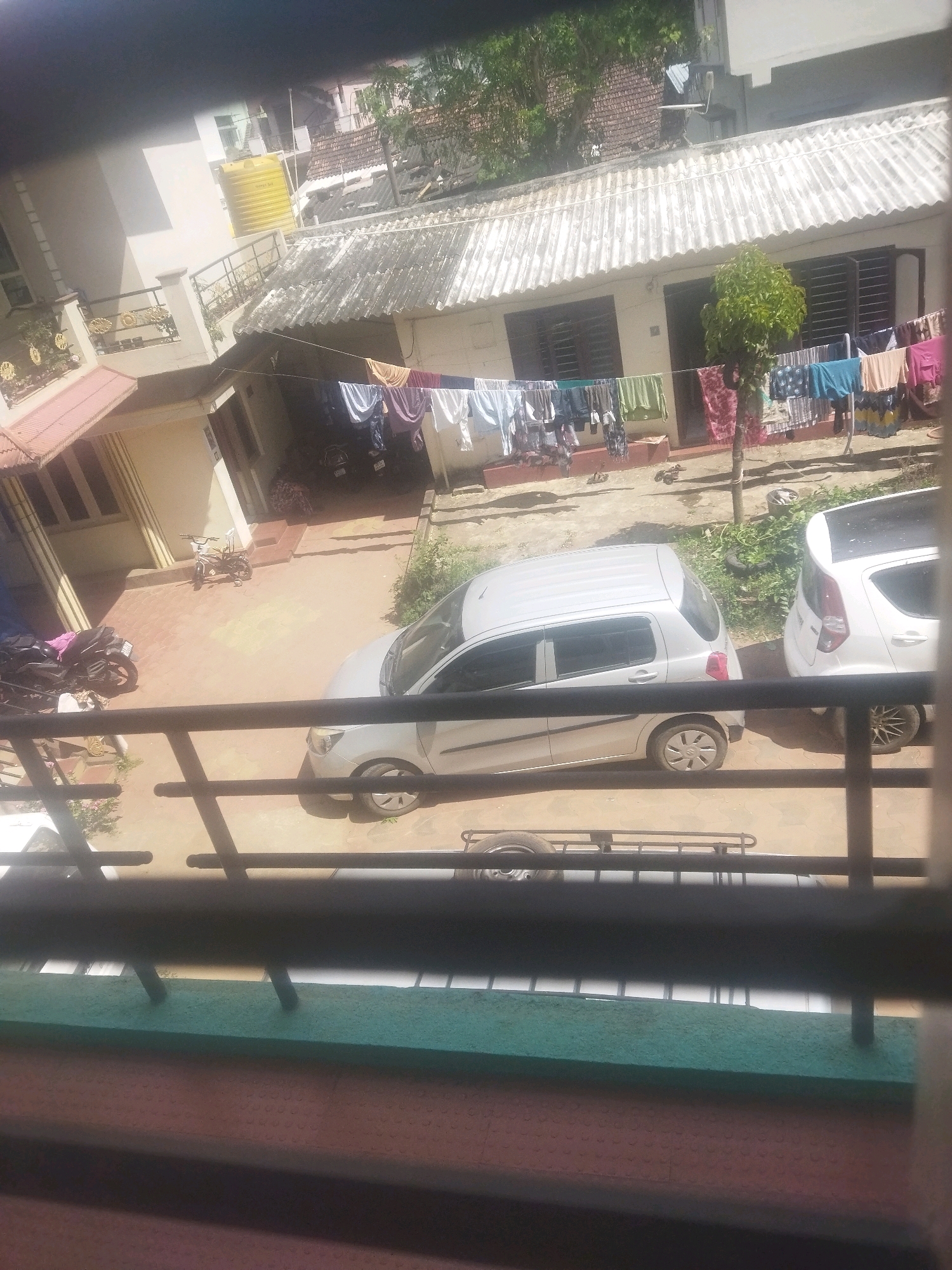 A high-angle, medium shot shows a courtyard with two cars, a motorcycle, and laundry hanging on a line in an urban environment. The courtyard is paved with interlocking bricks. To the left, a light-colored motorcycle is parked next to a blue tarp. A small, dark-colored motorcycle is parked further back in the courtyard, next to a small bicycle.
Two white cars are parked in the courtyard. The car on the left is a light silver hatchback with a dented rear bumper. The car on the right is a white hatchback with a black roof. A clothesline is strung across the courtyard, with various items of clothing hanging on it, including shirts, pants, and towels.
In the background, there are two-story buildings with tiled roofs. The building on the left has a yellow water tank on its roof. The building on the right has a row of windows and a tree growing in front of it. The scene appears to be in the daytime, with bright sunlight casting shadows on the ground. There are no people visible in the image. The overall impression is one of a residential courtyard in a densely populated urban area.