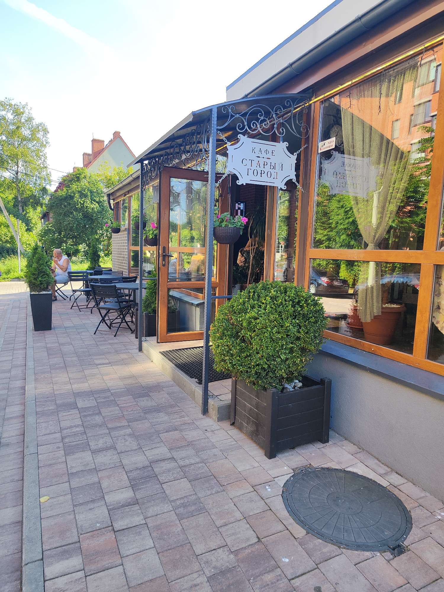 A street-level outdoor shot captures the entrance and exterior seating area of a cafe in Pionersky, Russia. The cafe features a glass-enclosed sunroom with wooden trim and a black wrought-iron awning above the entrance. A sign hanging from chains reads "КАФЕ СТАРЫЙ ГОРОД" (Cafe Old Town). Black metal tables and chairs are set up on a patterned brick patio. A lone man sits at one of the tables, looking at his phone. Several potted plants, including two tall conical evergreens and a large round bush in a rectangular planter, are placed around the entrance and seating area. The facade of the building is brick, and large windows reflect the surrounding trees and buildings. A manhole cover is visible on the brick sidewalk in the foreground. The sky is clear and blue, suggesting a sunny day.