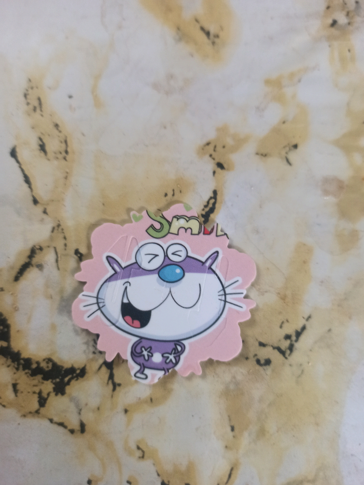 A sticker of a cartoon cat with large eyes, a wide smile, and a blue nose is placed on a marbled surface. The cat has purple accents on its head and wears a purple bow with white Xs on its chest. Above the cat's head, the word "SMILE" is written in green and yellow letters. The background appears to be a countertop or table with a beige and white marbled pattern, featuring dark brown and black irregular markings. The lighting suggests an indoor setting. The image does not contain any information or context related to Machraa El Ain, Morocco.