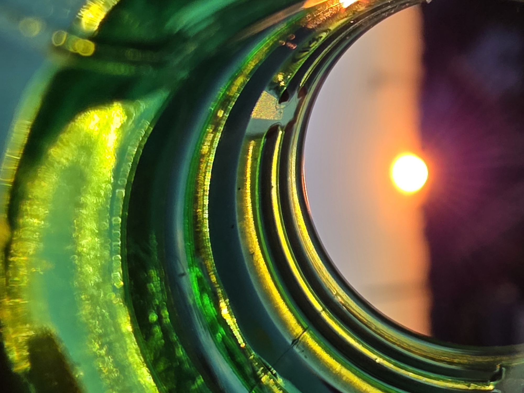 The image shows a close-up view through the opening of a green glass bottle. The bottle's rim is heavily textured with concentric rings and reflective surfaces. The light refracting through the glass creates bright green and yellow highlights. Looking through the bottle, a bright, round object, likely the sun, is visible against a blurred background of orange, white, and purple hues, potentially depicting the sky at sunset. There is no context to definitively place this scene in Chaikovsky, Russia. No people or other specific objects besides the bottle and the light source are discernible. The image captures the interaction of light and glass to create an abstract, visually arresting composition.