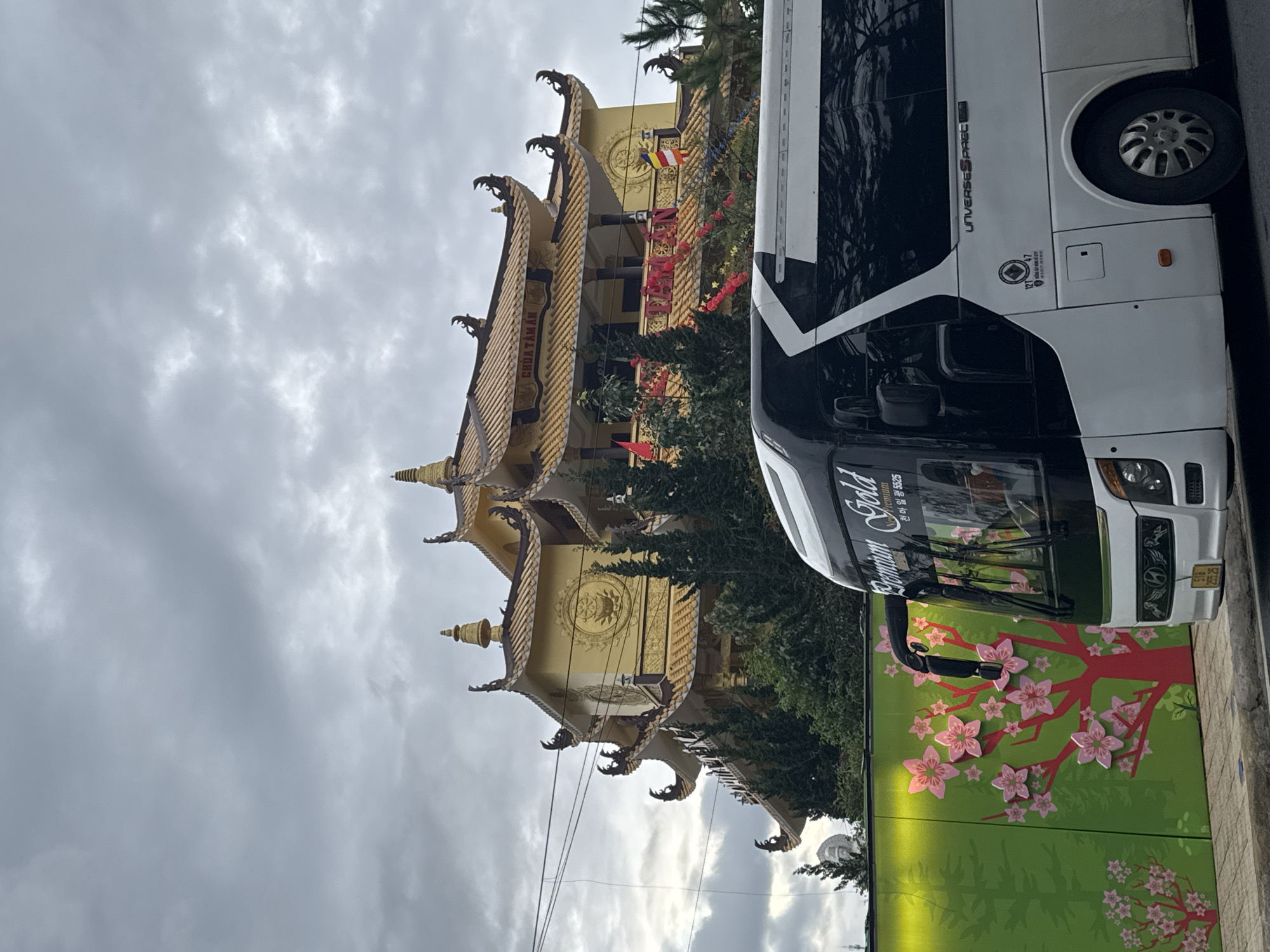 The image depicts the Chua Tam An temple in Da Lat, Vietnam, from a street-level perspective. The temple is a multi-tiered golden structure with ornate, stylized roof edges. A white and black bus occupies the right side of the frame, partially obscuring the temple. The bus has the words "Premium Gold" written on the side, along with Korean text. A green barrier with a cherry blossom design is positioned in front of the bus. The sky is overcast with gray clouds. The temple is partially obscured by trees. A Vietnamese Buddhist flag is visible on the temple.
