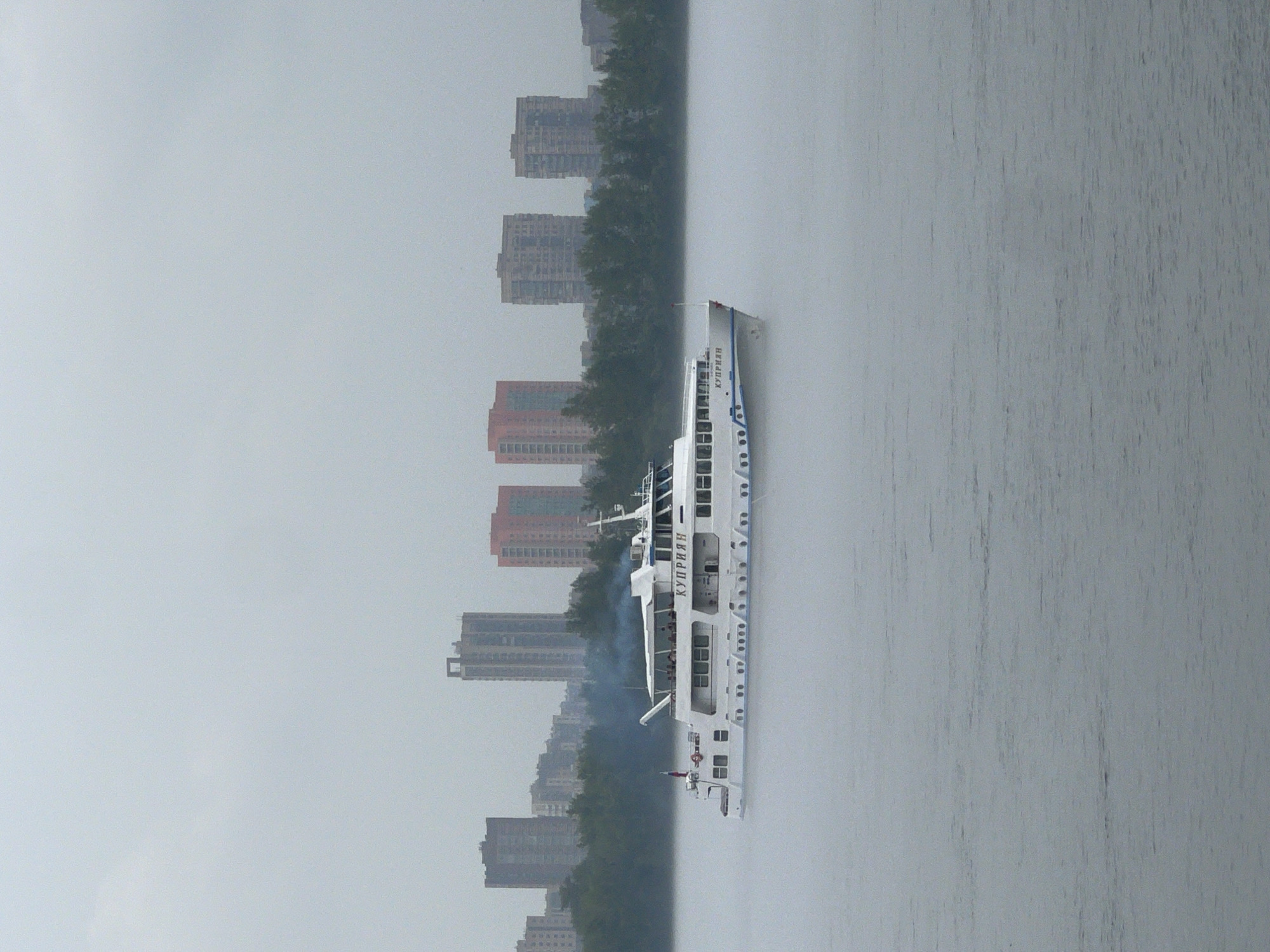 A white passenger ship named "КУПРИЯН" sails on a river under a cloudy sky. The ship is in the foreground, and a hazy cityscape with several tall buildings and trees is visible in the background. The city is located in Krasnoyarsk, Russia. The river is calm, with slight ripples on the surface. The ship has several decks and windows, and people can be seen on the upper deck. Smoke is emanating from the ship's smokestack. The overall atmosphere is somewhat muted due to the overcast weather and mist.