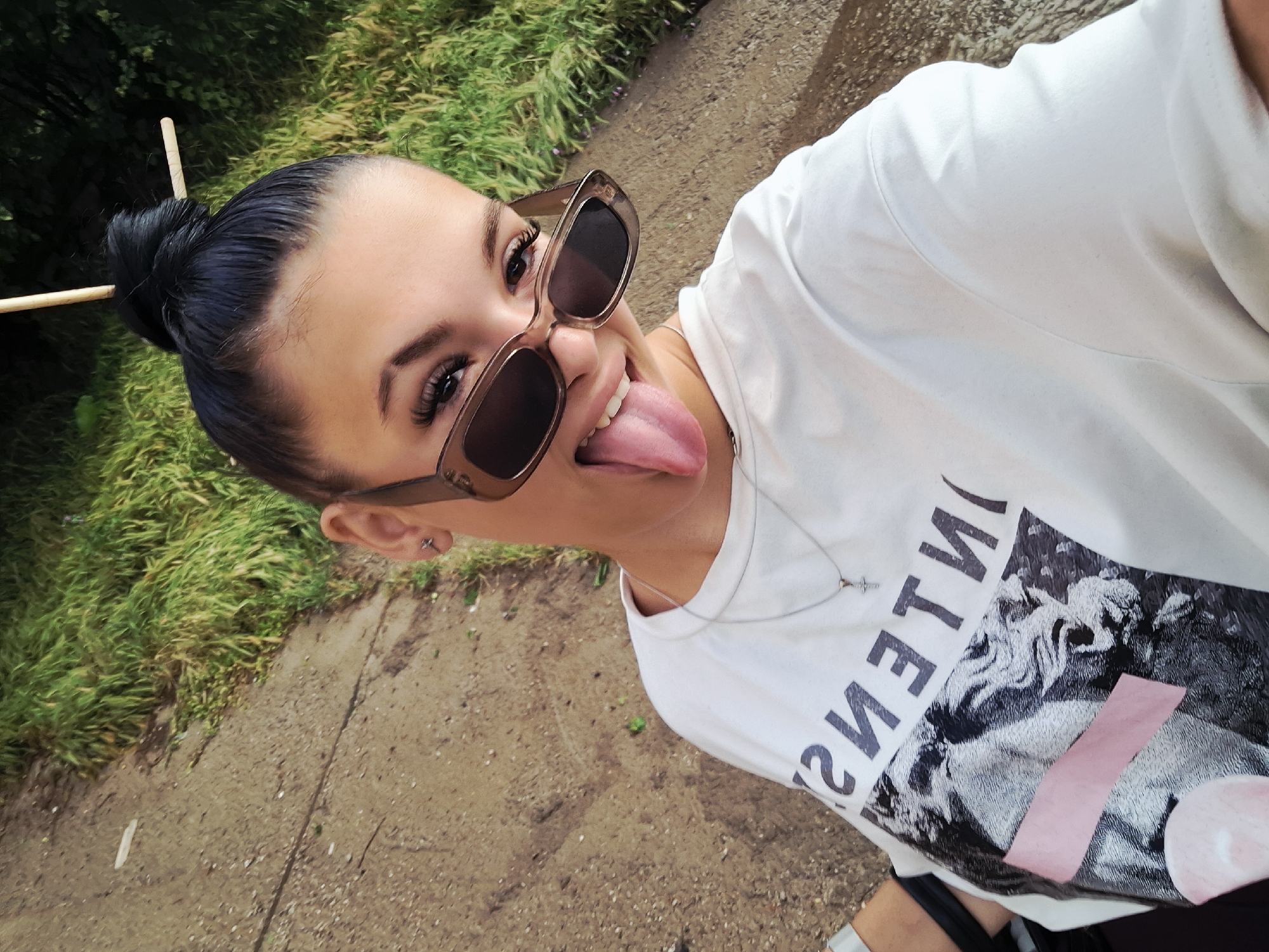 A young female, appearing to be an adult, is depicted in a close-up selfie taken outdoors in Krapets, Bulgaria. She is looking directly at the camera with a playful expression, her tongue extended. Her dark hair is styled in a high bun, secured with two wooden sticks. She is wearing translucent brown-framed sunglasses with dark lenses, positioned slightly askew on her face. Her visible right eye (left from the viewer's perspective) features prominent eyelashes and eyeliner. She wears a white short-sleeved t-shirt with a mirrored black text graphic reading "ESNETNI" (INTENSE) above a black and white image of a classical sculpture, overlaid with a pink horizontal rectangle and a subtle pink circular element. A thin silver-colored chain necklace with a small cross pendant is visible around her neck. Her right ear has a small stud earring. The background consists of a light brown, textured ground, possibly a dirt or gravel path, and a dense patch of green foliage on the right and top. The scene is illuminated by natural daylight.